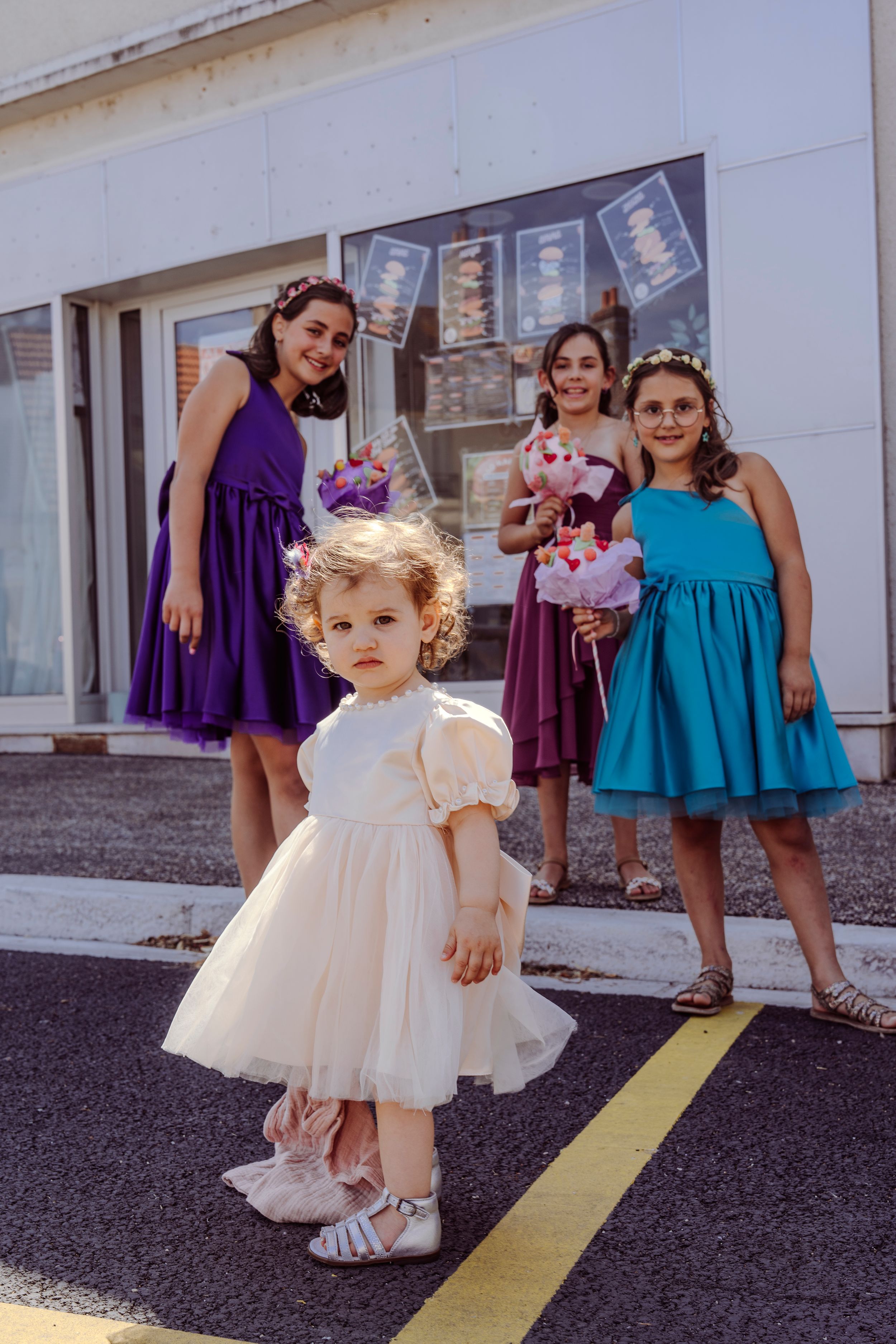 A young child in a white tulle dress twirls and plays with others wearing purple and teal dresses outdoors.