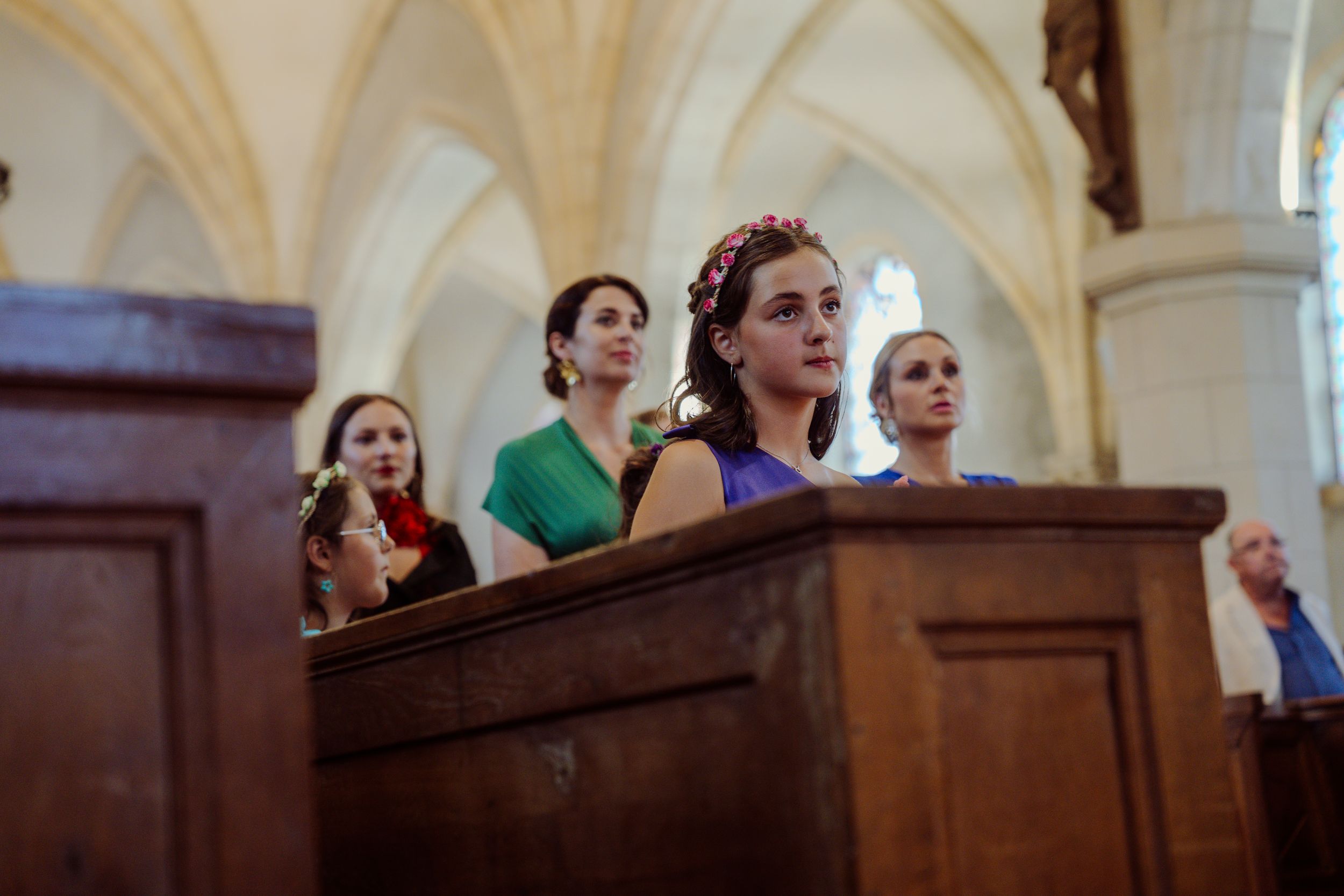 Wedding attendees stand in a wooden church pew during a ceremony in a vaulted cathedral interior.