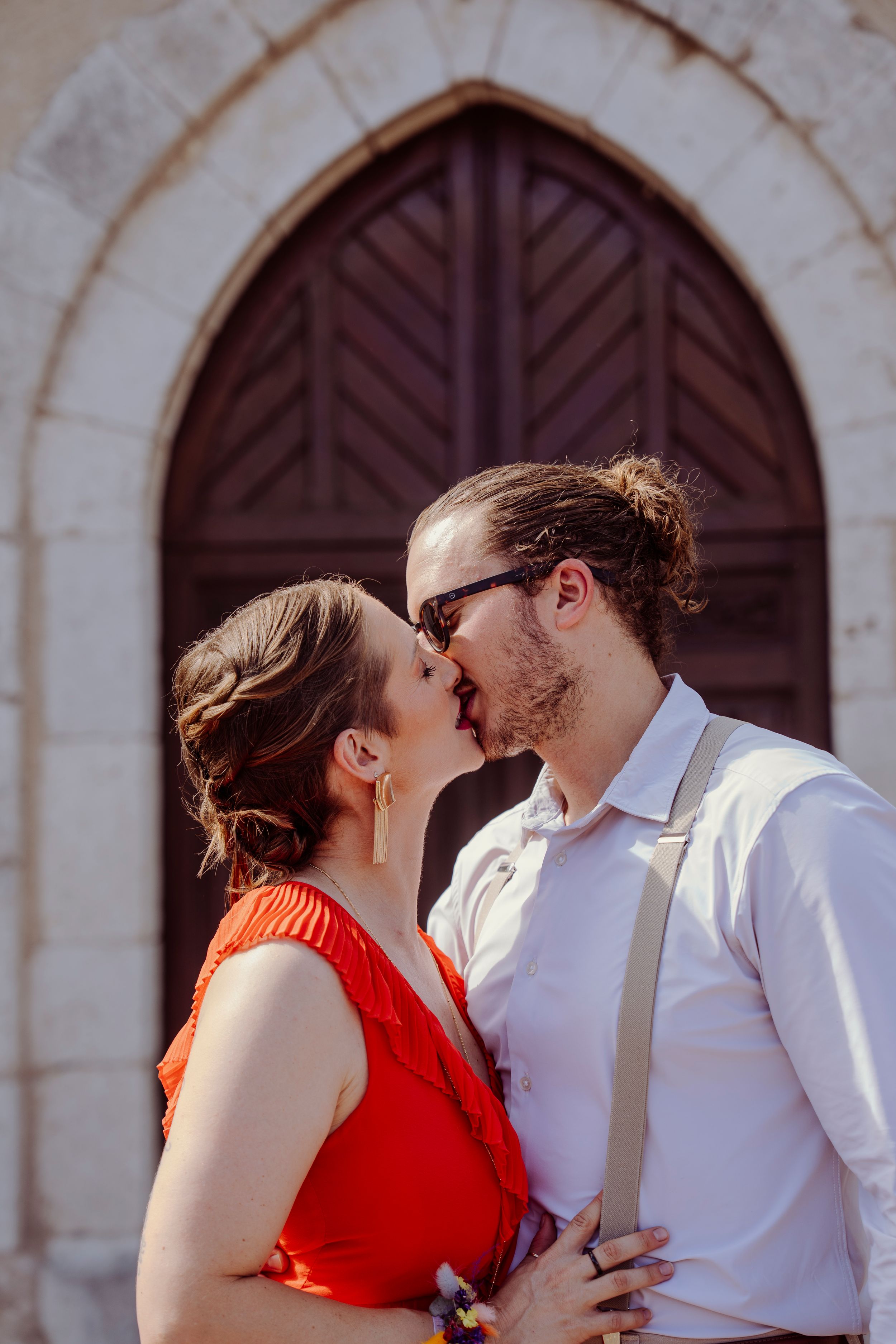 A romantic couple shares intimate kisses in front of an arched stone wall with wooden doors.