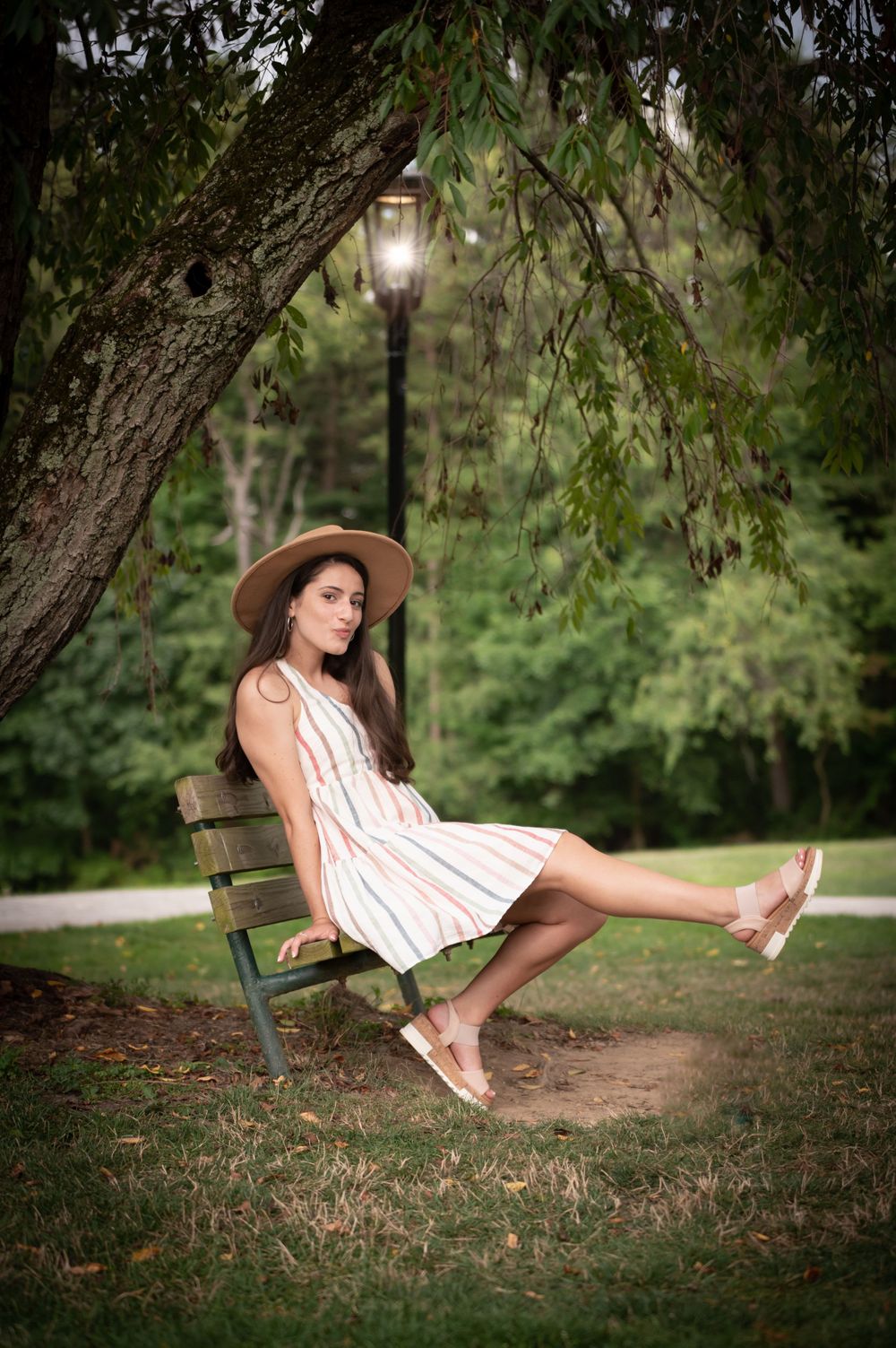 A person in a white sundress and hat relaxes on a park bench under a tree with a glowing lamp post.