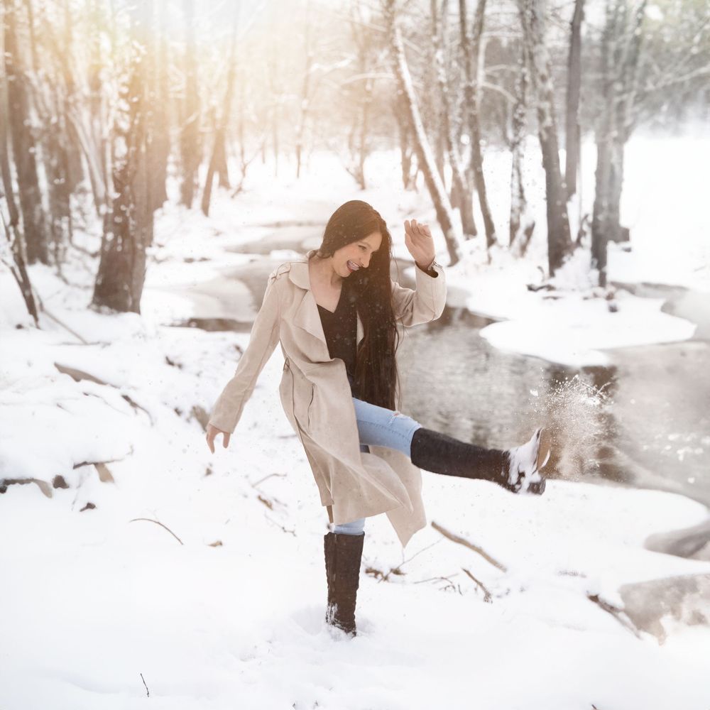 A person in a beige coat walks through a snowy winter forest scene with sunlight filtering through trees.