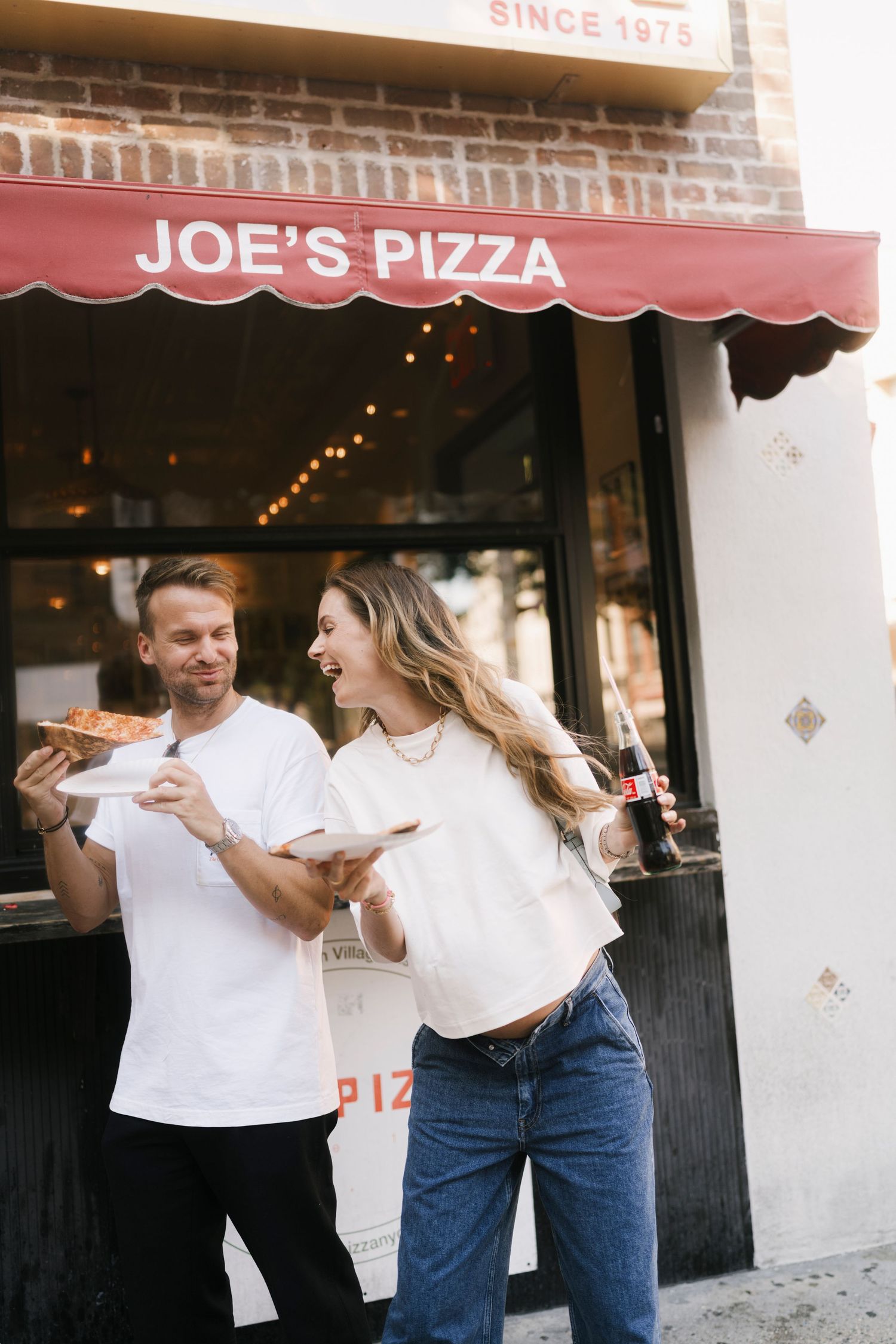 Two people enjoying pizza and laughing together outside Joe's Pizza restaurant storefront with a red awning.