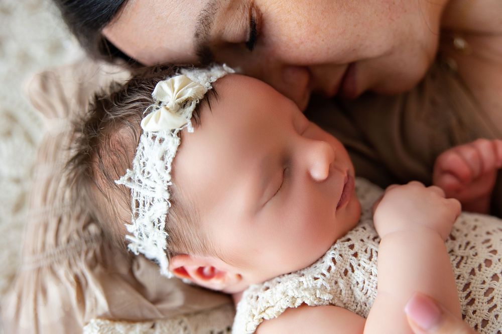 A sleeping baby wearing a white lace outfit rests peacefully on soft beige bedding.
