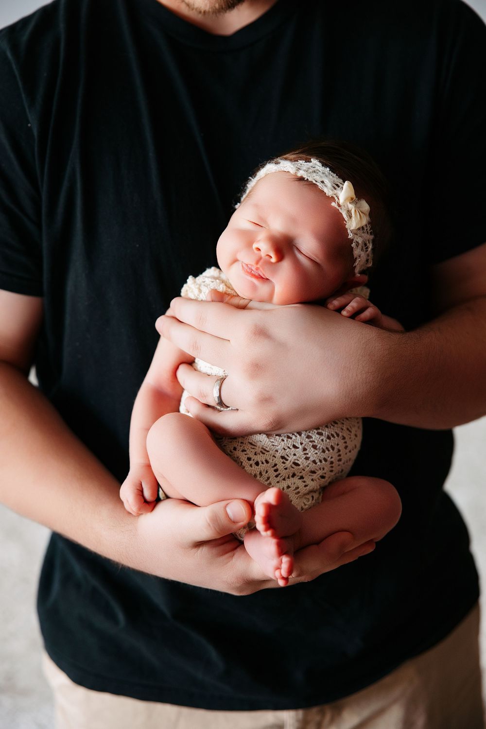 A sleeping newborn baby wearing a delicate lace headband and knit diaper cover is cradled gently in someone's hands.