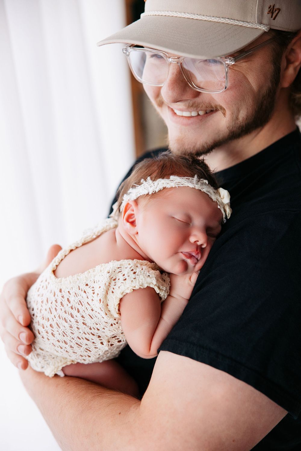 A sleeping baby in a white crochet dress rests peacefully against someone wearing a black shirt.