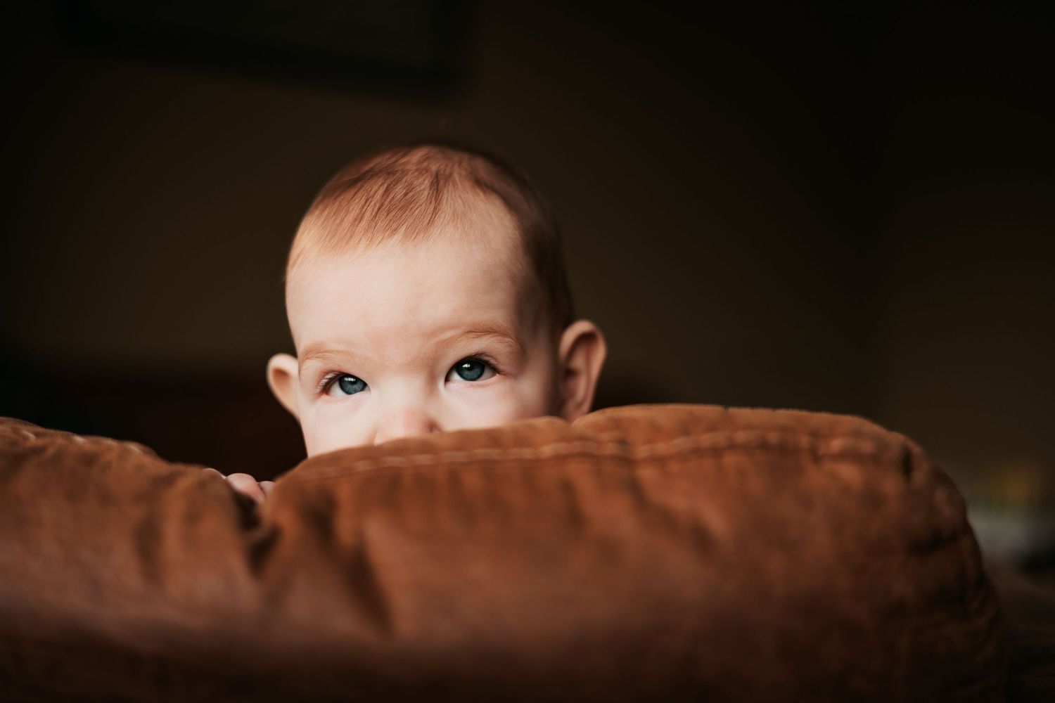 Curious baby peeking over a brown cushion with bright eyes in a cozy home | In-Home Family Photos in Westminster, CO