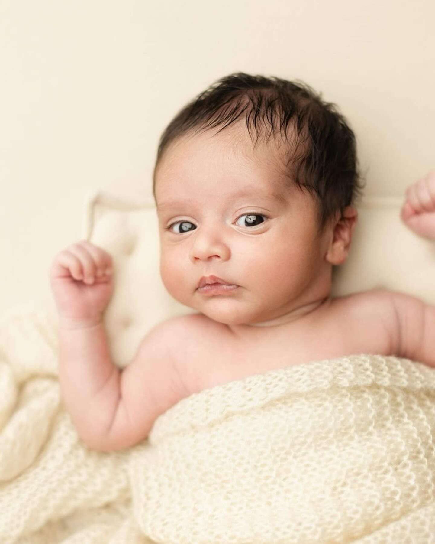 A small baby wrapped in a cream-colored knit blanket lying on white bedding in soft natural light.