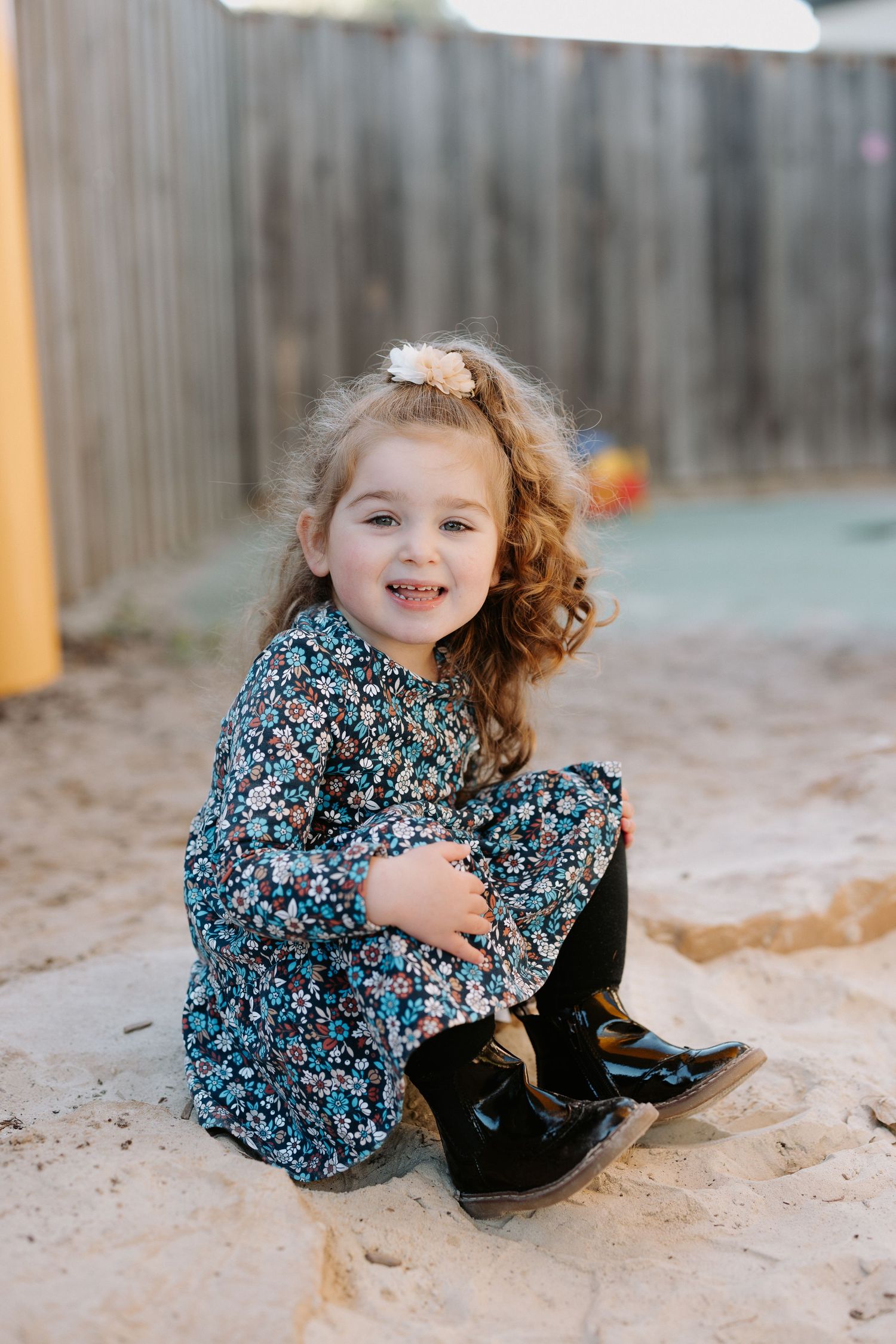 A child in a floral dress crouches down and smiles while playing in a sandy area outdoors.