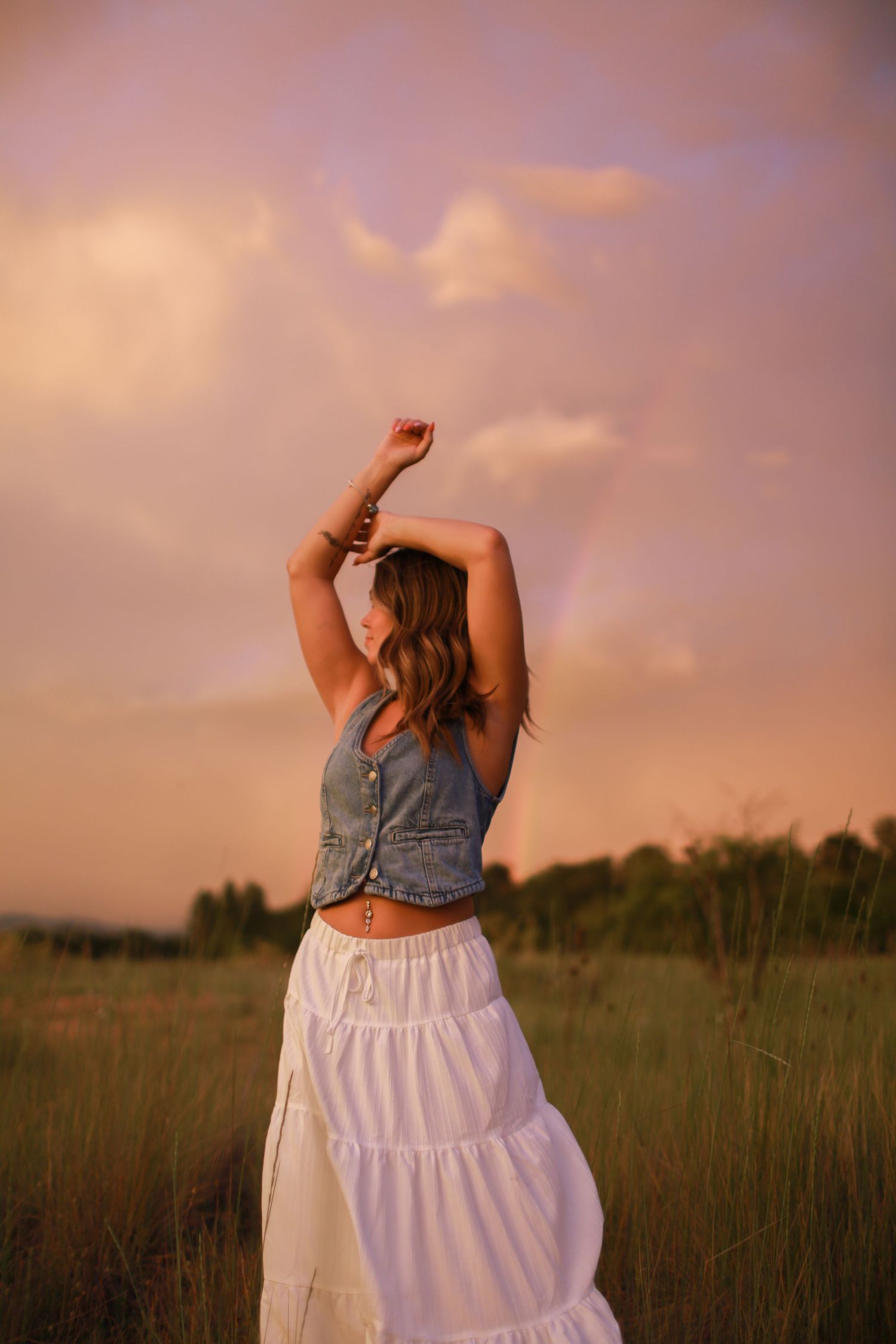 A figure in a denim vest and white skirt stands in a golden field at sunset, creating a dreamy silhouette.