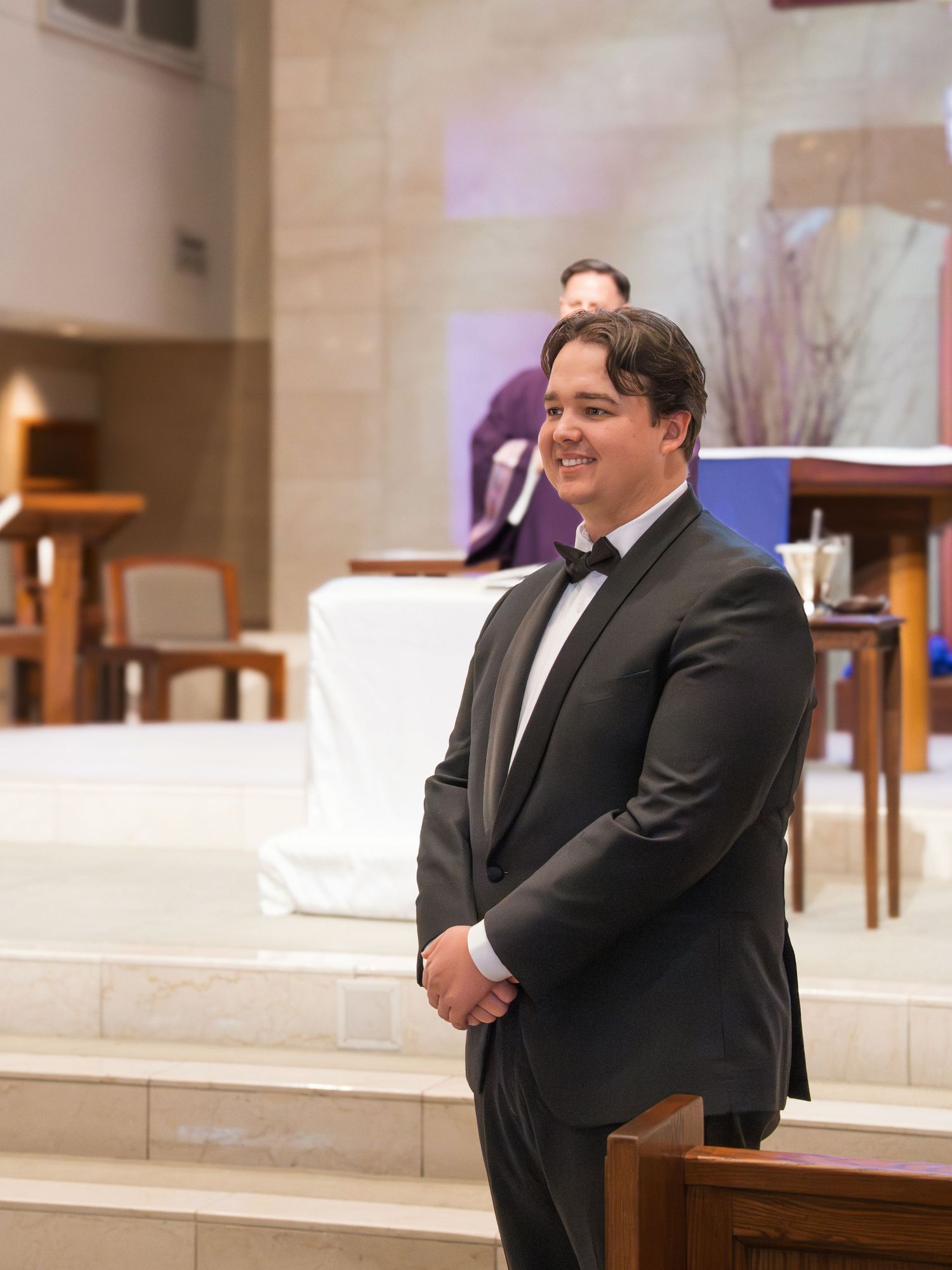 A groom in a black suit smiles during a wedding ceremony in a church setting.
