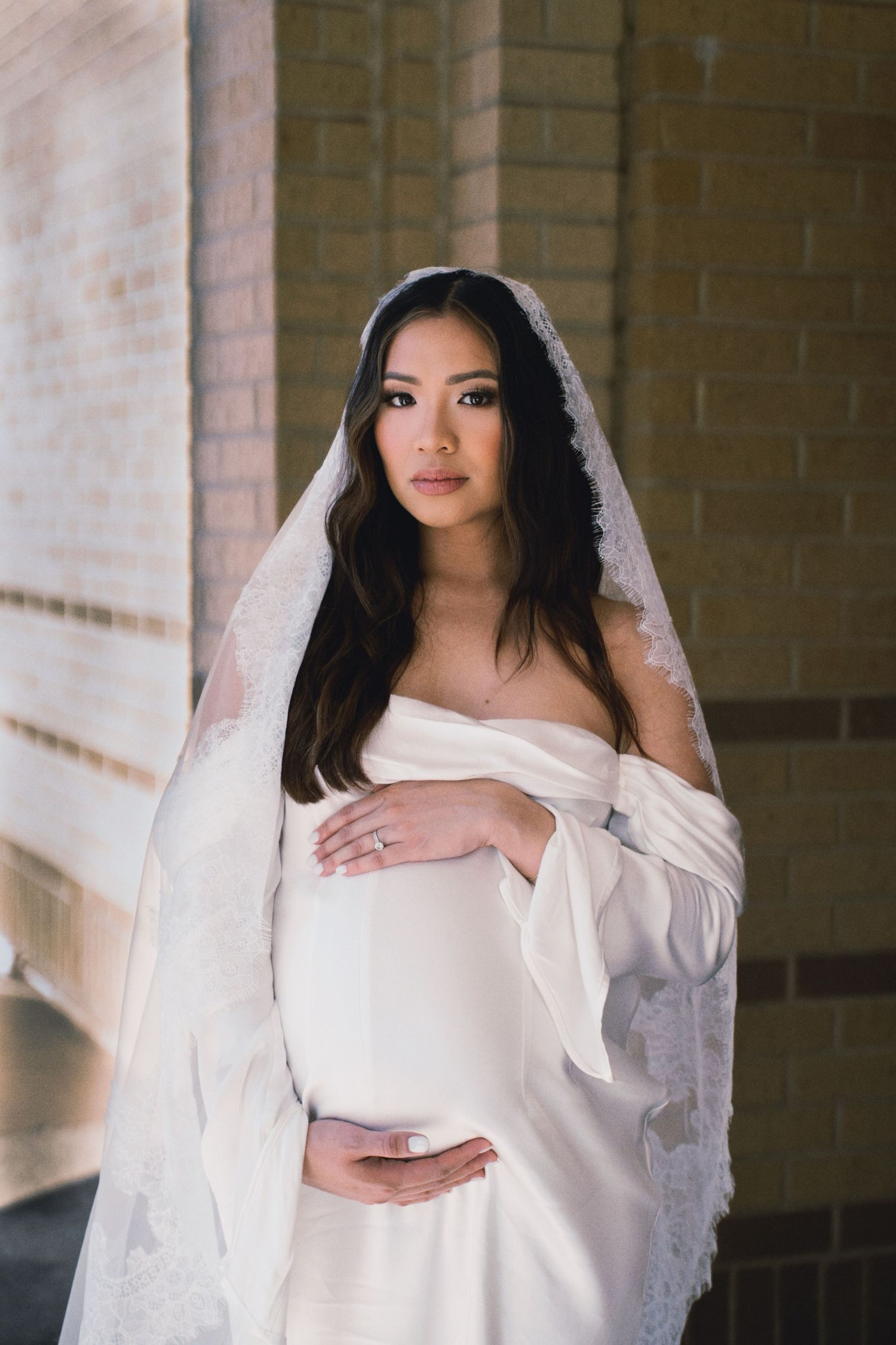 Elegant maternity portrait in white dress and veil against brick wall with soft natural lighting.