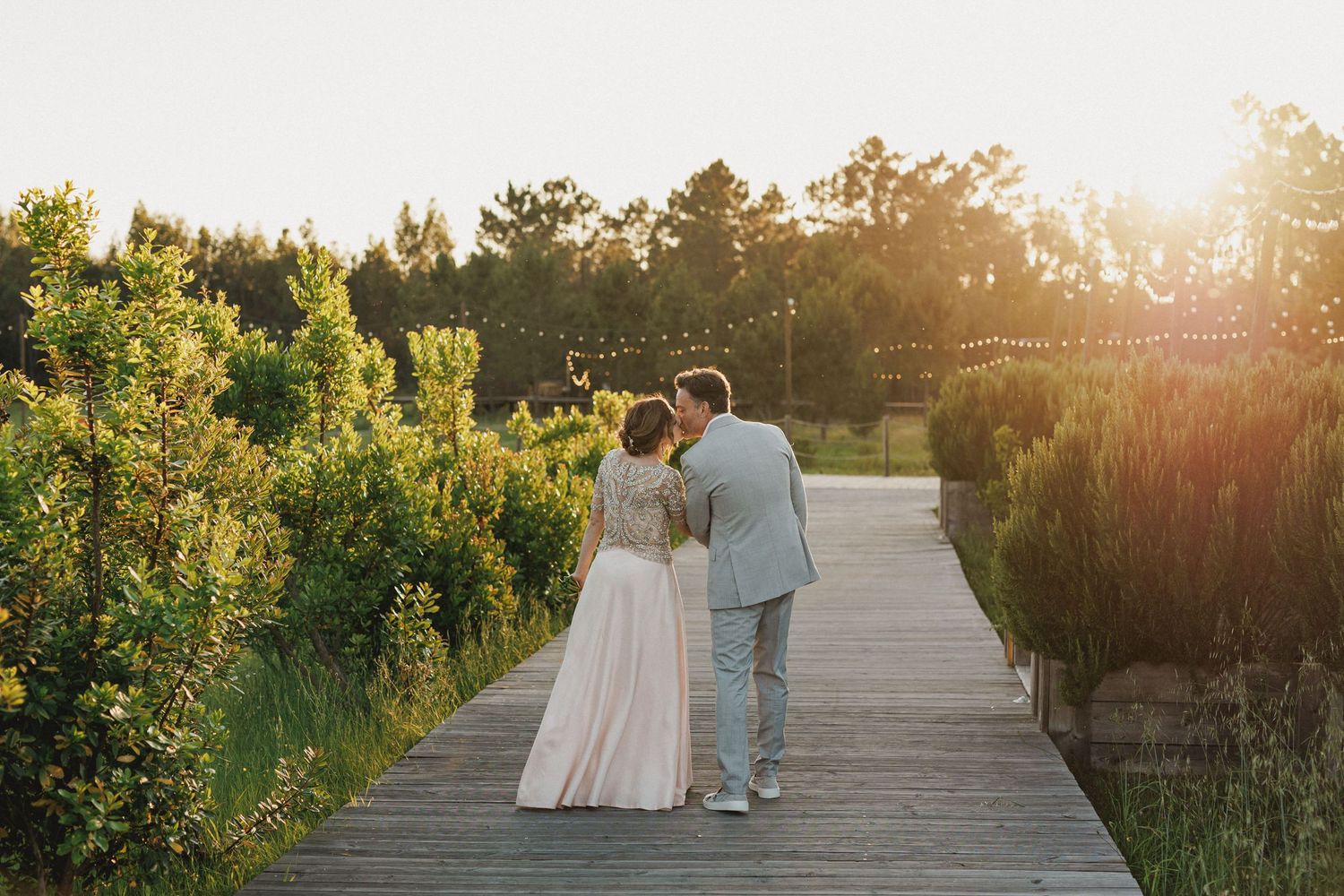 A couple walks on a wooden boardwalk through natural vegetation at golden sunset hour at their wedding at Craveiral Farmhouse in alentejo
