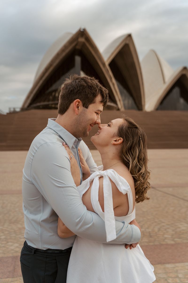 A romantic couple hugs and snuggles in front of the iconic Sydney Opera House on a cloudy day during their engagement photoshoot