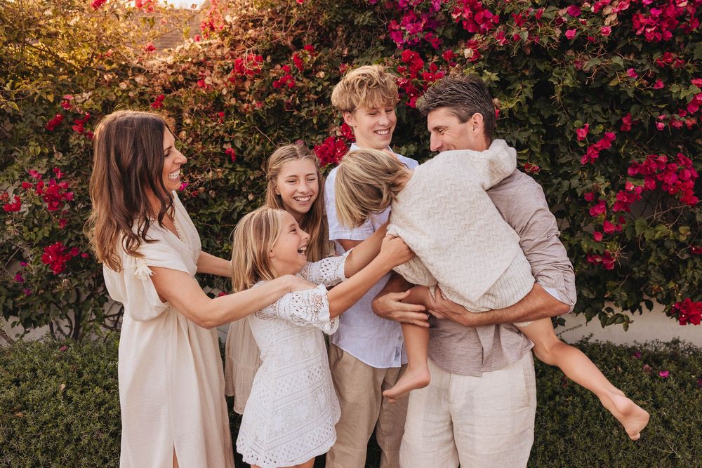 A family wearing neutral colors poses together in front of vibrant red bougainvillea flowers in a garden setting.