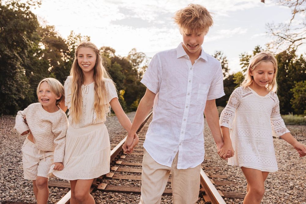 A family wearing white and beige outfits poses together outdoors during golden hour on a gravel path.