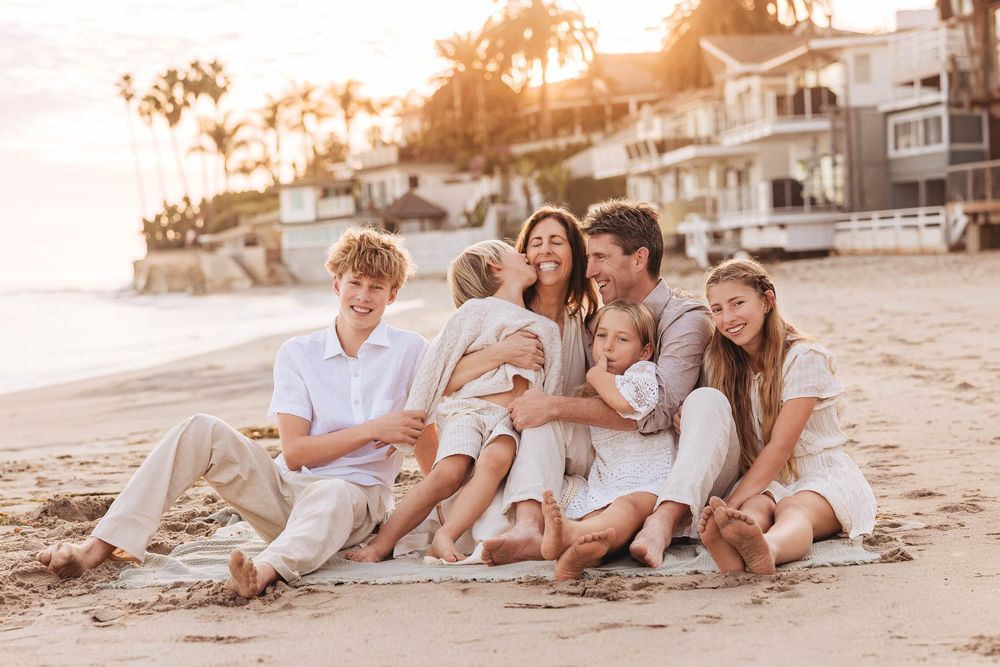 A family wearing white clothing relaxes together on a sandy beach at sunset.