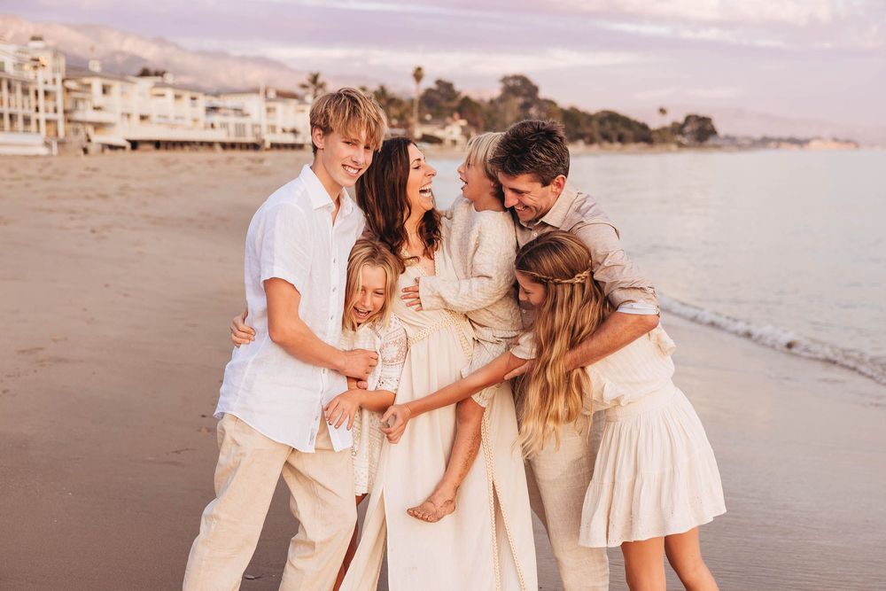 A family dressed in white embraces together on a beach at sunset with coastal buildings in the background.