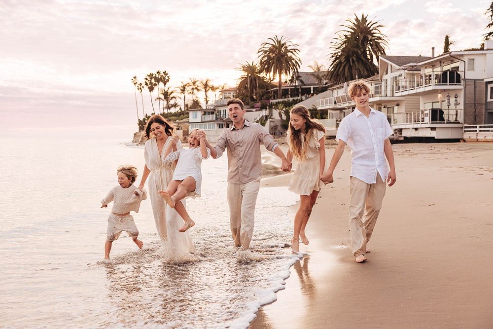 A group walks along a beach at sunset, splashing in the waves with palm trees and coastal homes in the background.