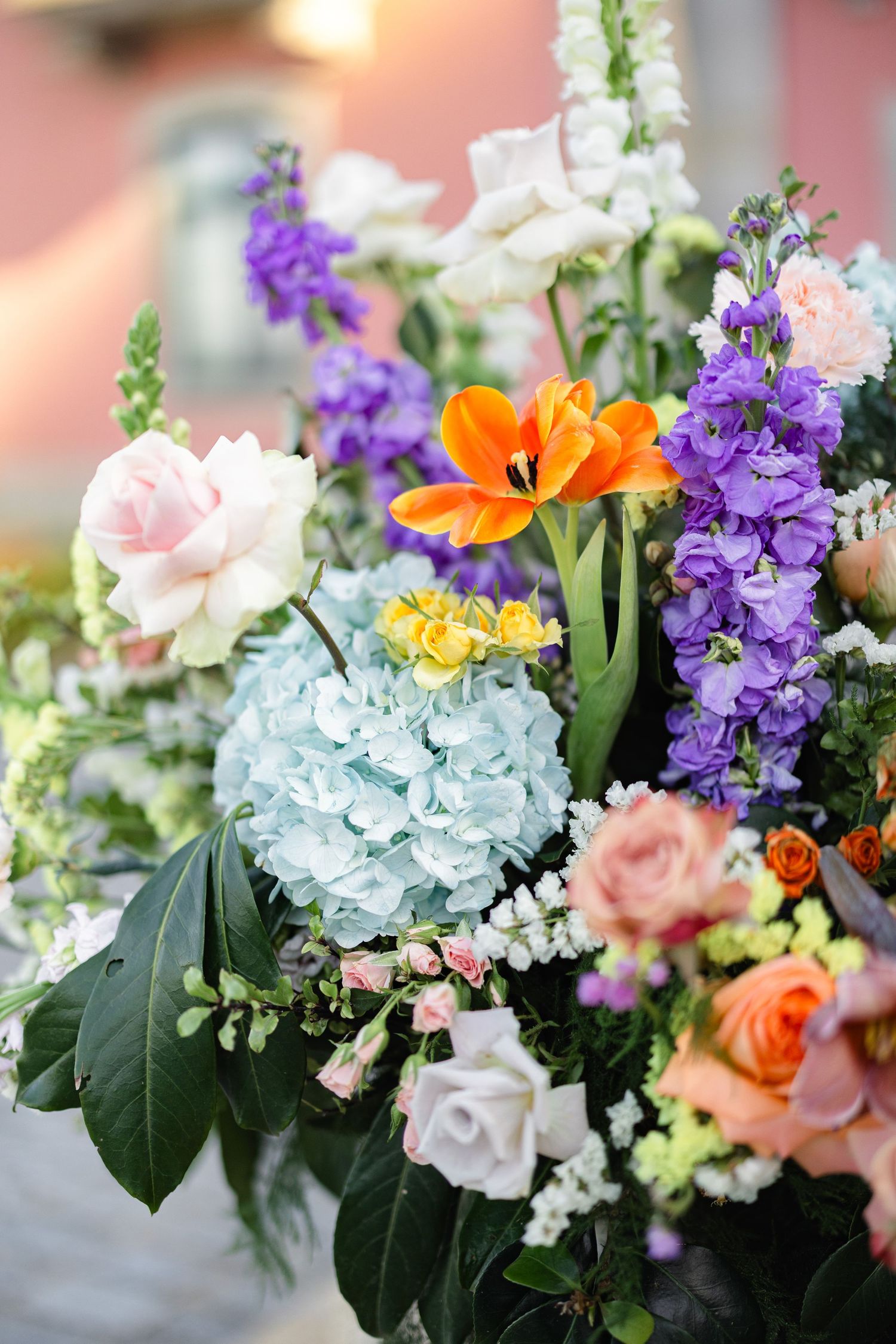 Colorful floral arrangement featuring orange tulips, purple delphiniums, blue hydrangeas, and pink roses in a lush bouquet.