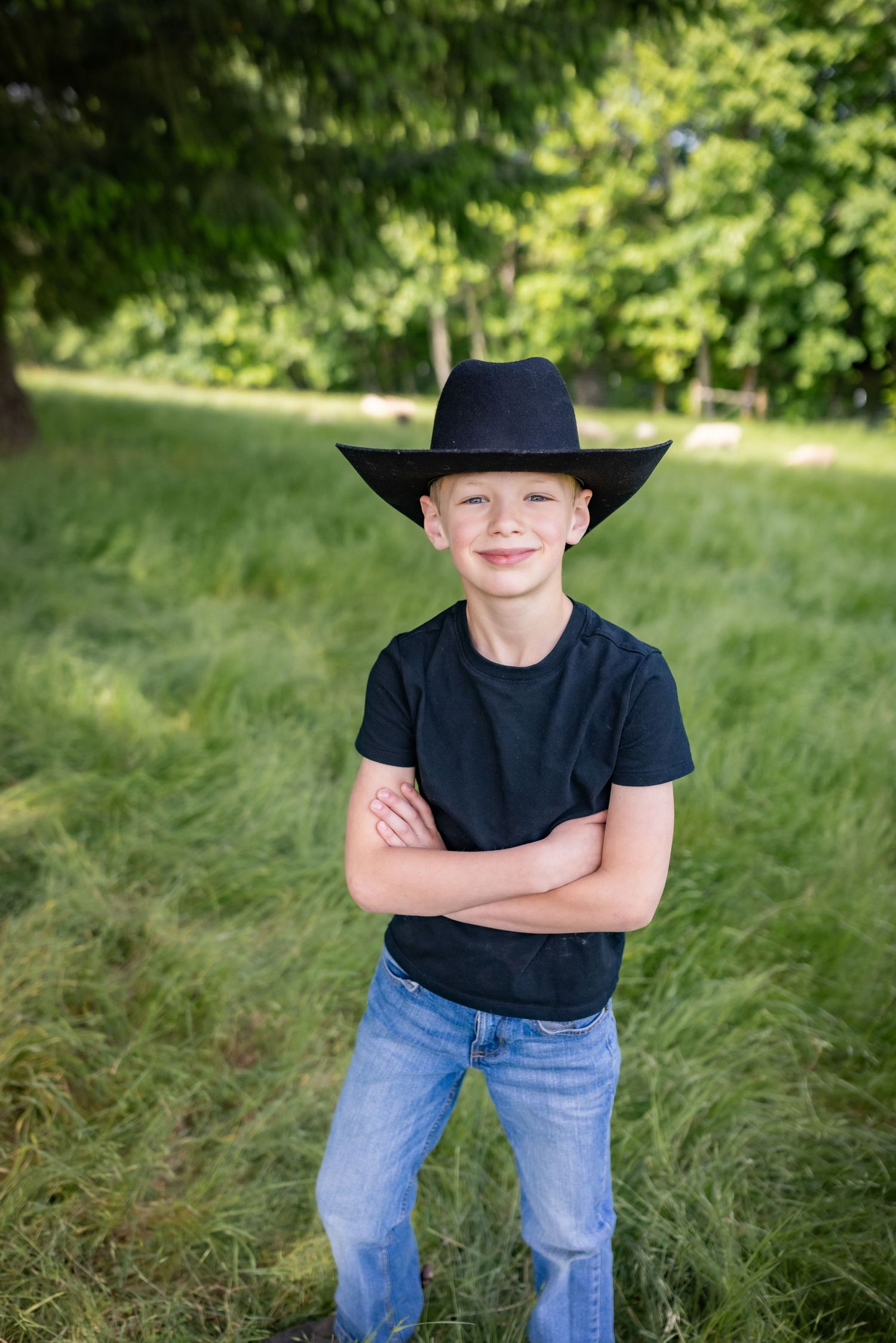 Confident boy in black cowboy hat and t-shirt stands with arms crossed in a green field.