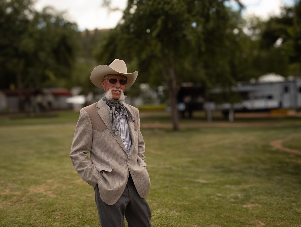 A person in western attire stands in a grassy field wearing a cowboy hat and beige jacket.