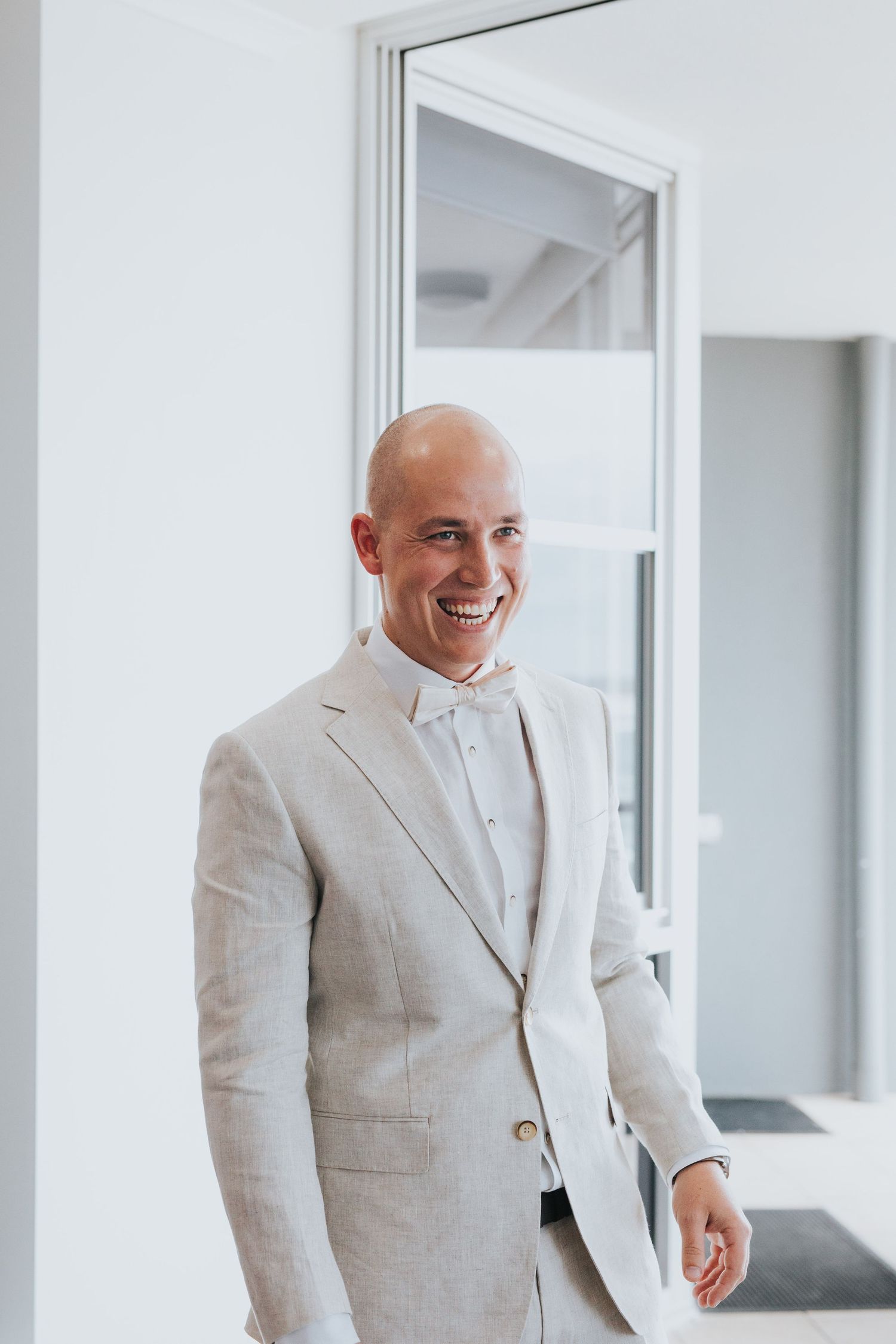 A person in a light beige suit laughs joyfully while standing in a bright modern interior space.