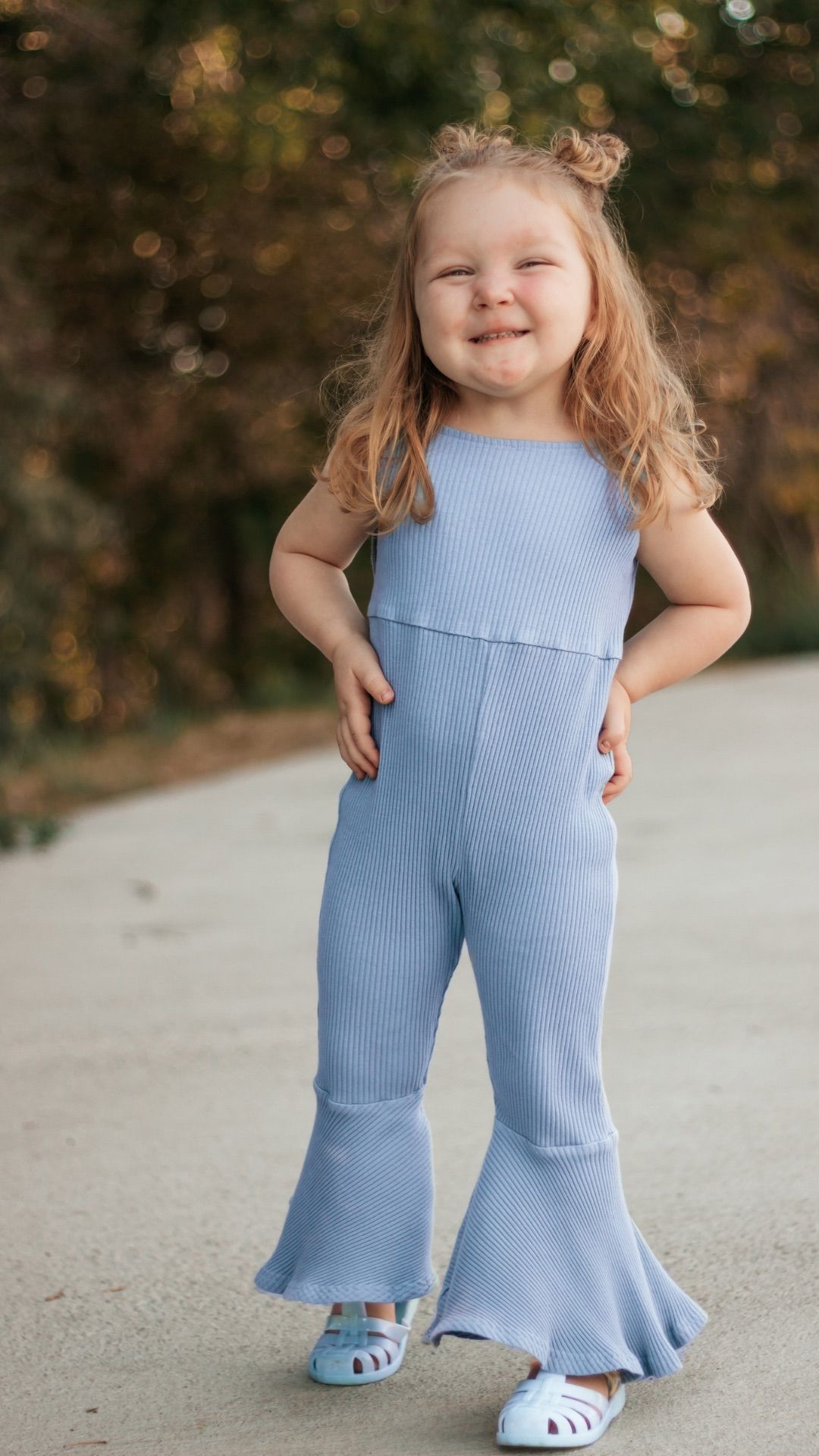 Smiling toddler girl in light blue bell-bottom jumpsuit posing outdoors with hands on hips | childhood photographer Westminster Colorado