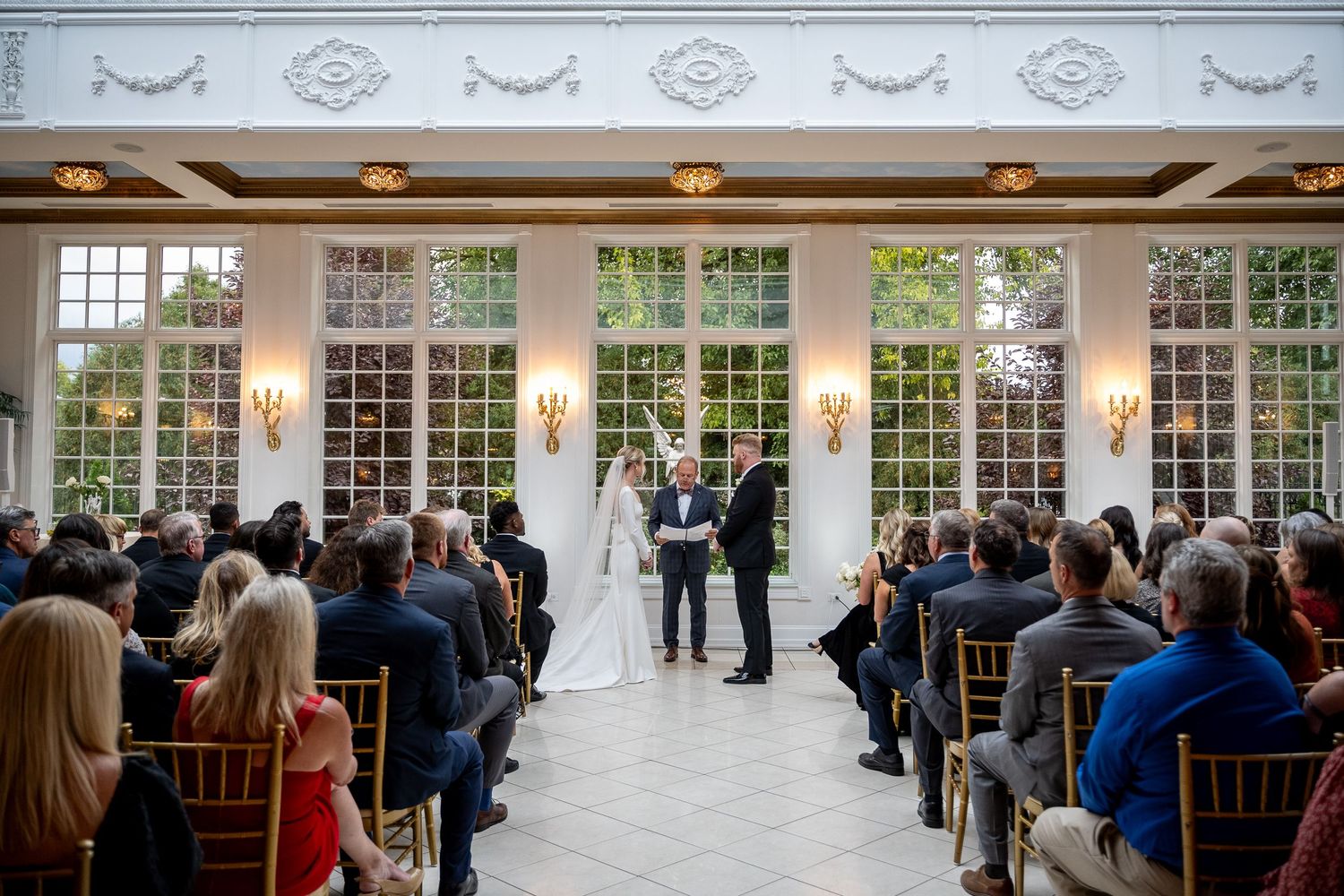 Elegant wedding ceremony setup with white columns, ornate ceiling, and guests seated in gold chairs in a bright ballroom.