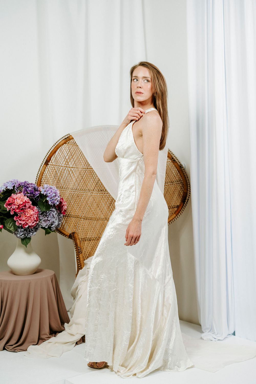 Elegant bridal photoshoot featuring a cream satin gown and wicker peacock chair with pink and purple floral arrangements.