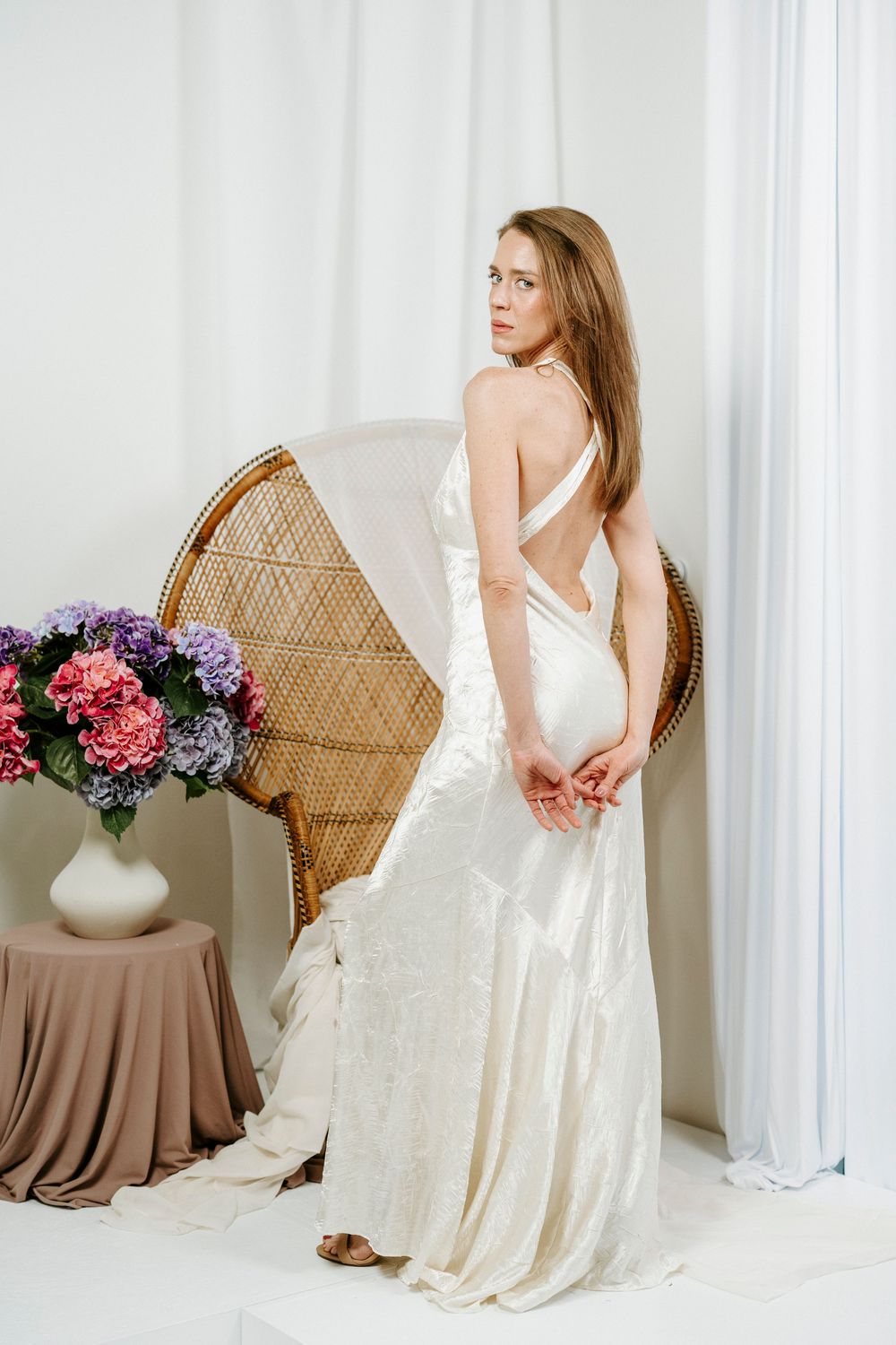 Romantic bridal portrait session with flowing ivory dress and rattan chair against white curtain backdrop.