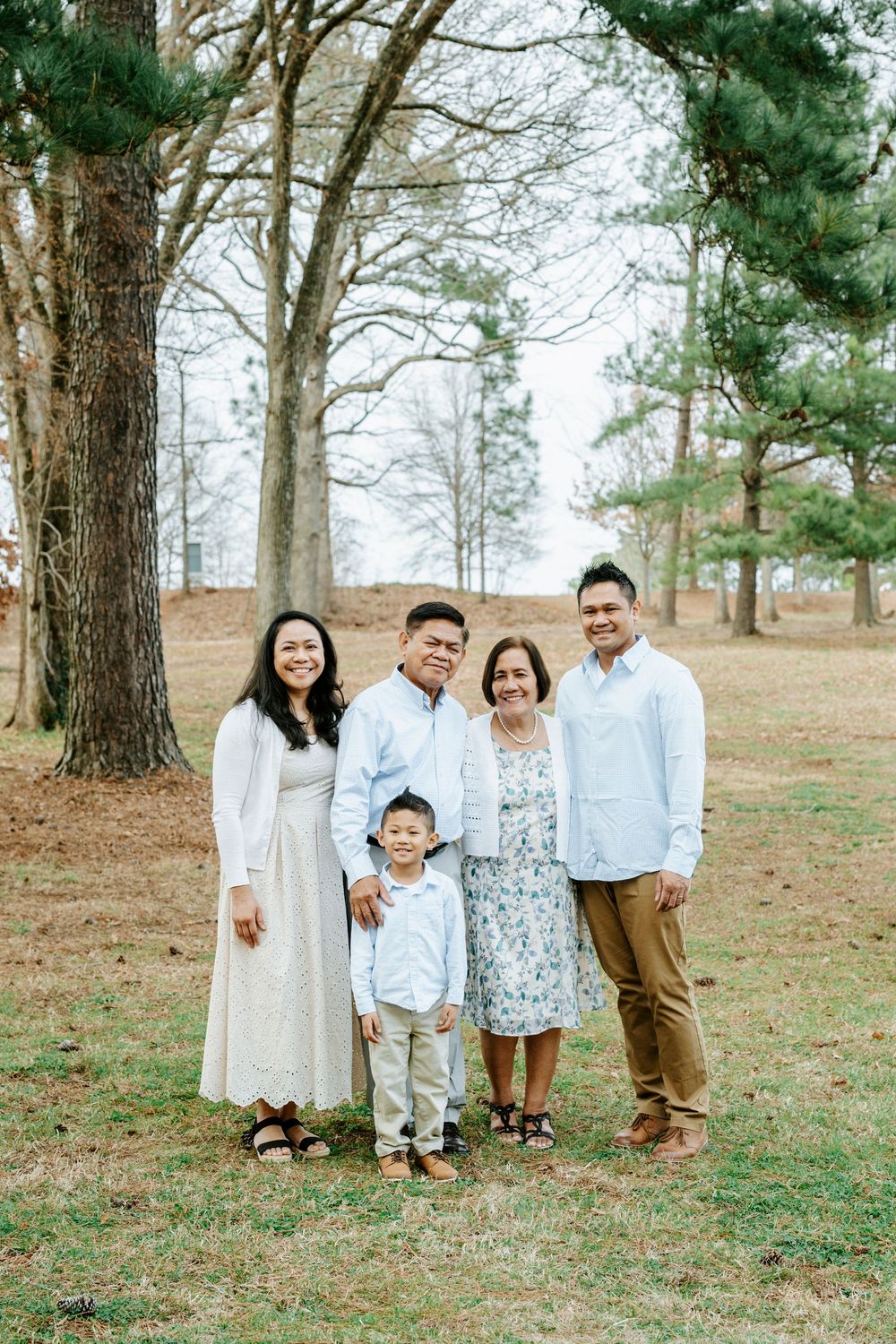 A family group wearing coordinated white and neutral outfits poses together on a grassy hill among tall pine trees.