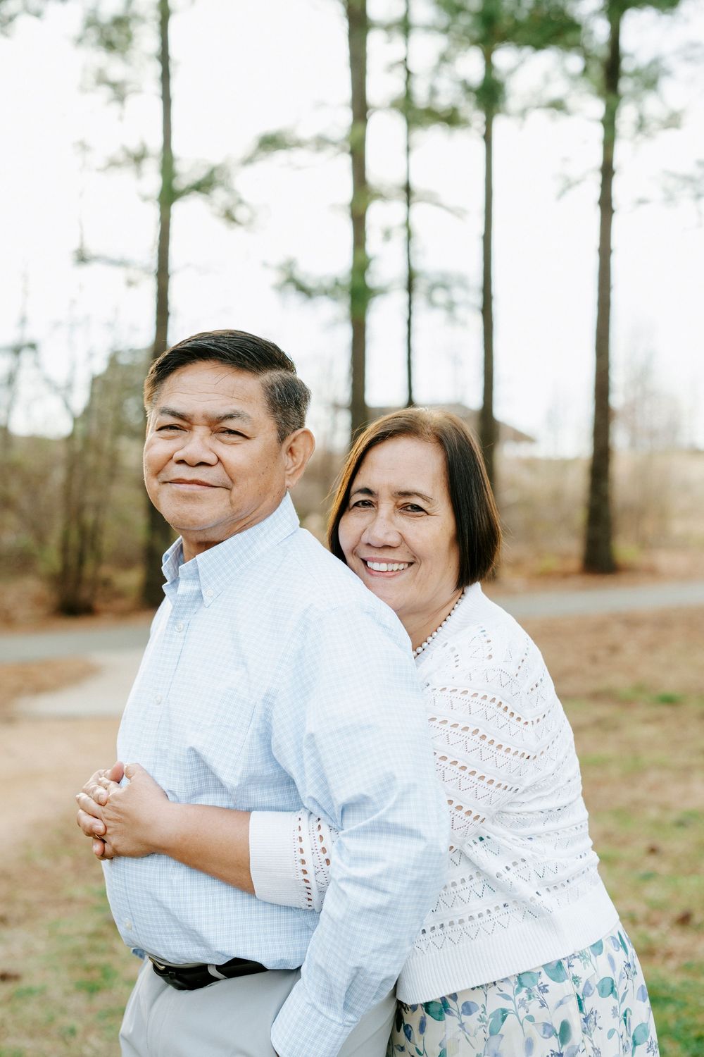 Couple in white outfits embracing outdoors with tall pine trees in the background.