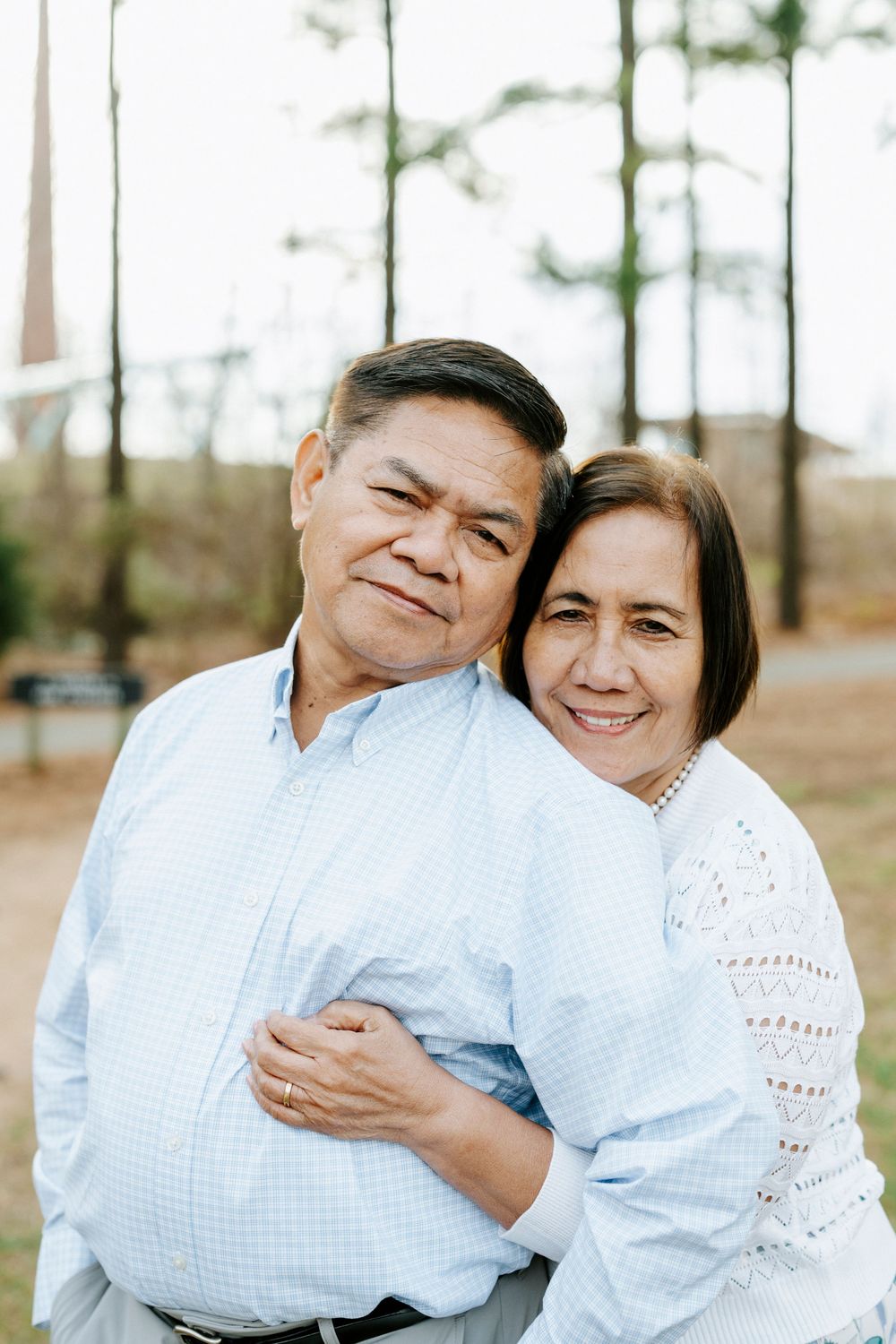 A loving couple wearing white outfits embrace while smiling outdoors in a pine forest setting.