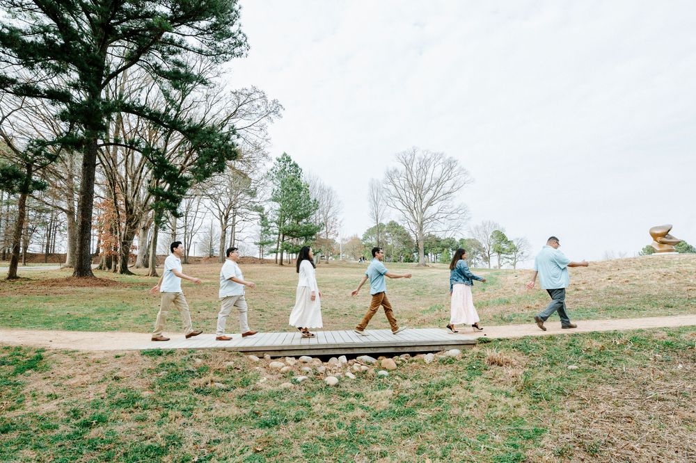 A line of people walk single file along a winding path through a scenic park with tall pine trees on an overcast day.