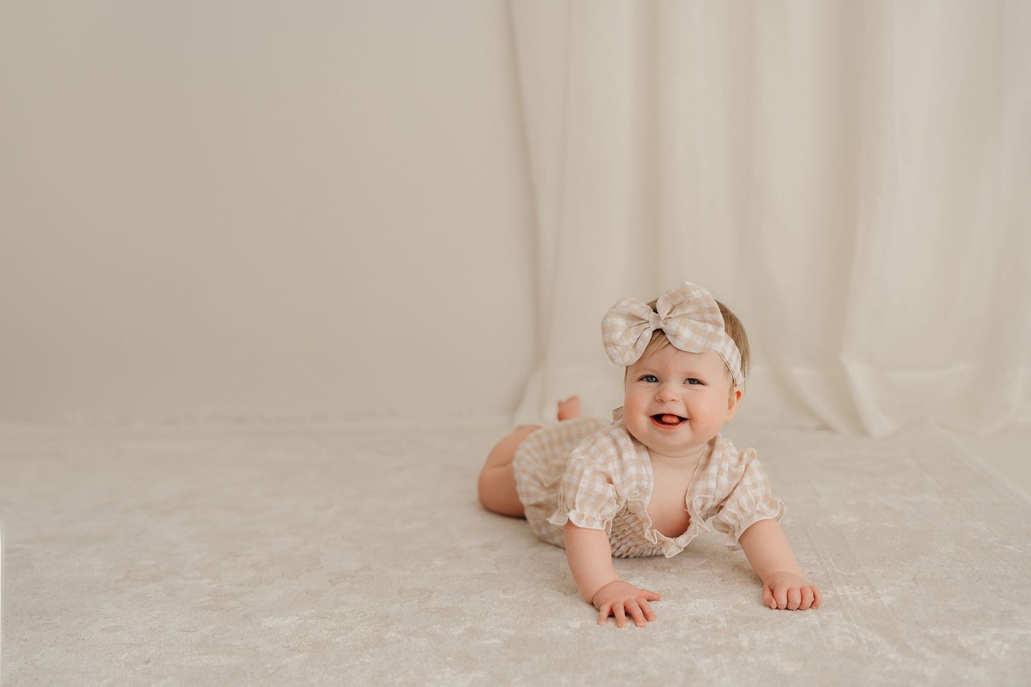 A small baby wearing a white bow headband lays on a light colored blanket with a sweet smile.