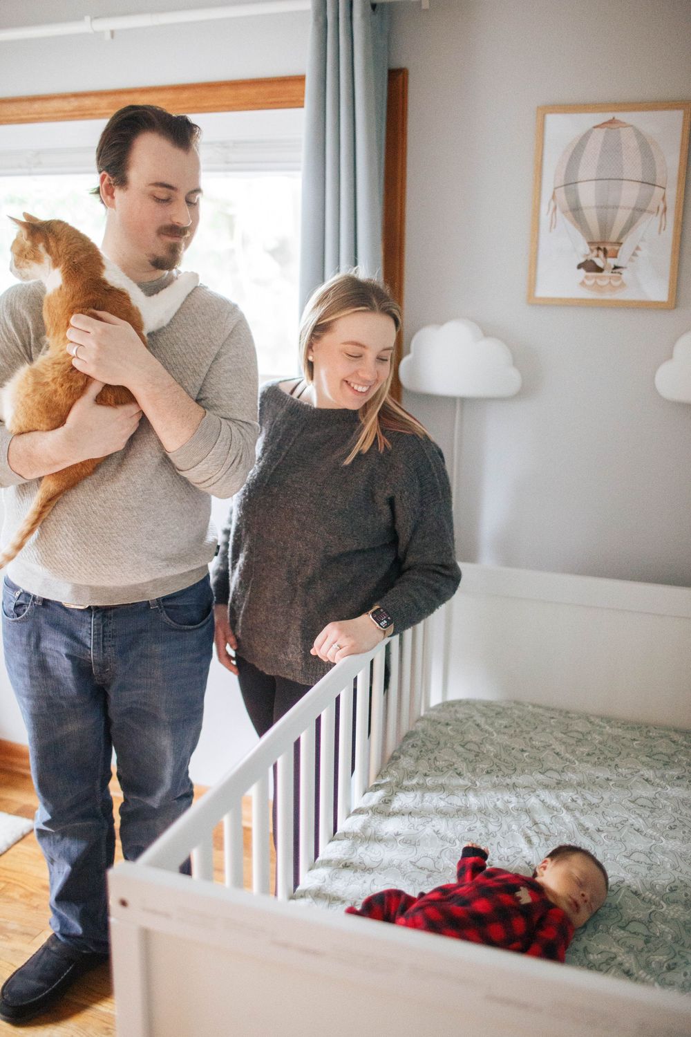 A mother and father hold their pet cat, as they look at their baby in the crib during an in-home lifestyle newborn session