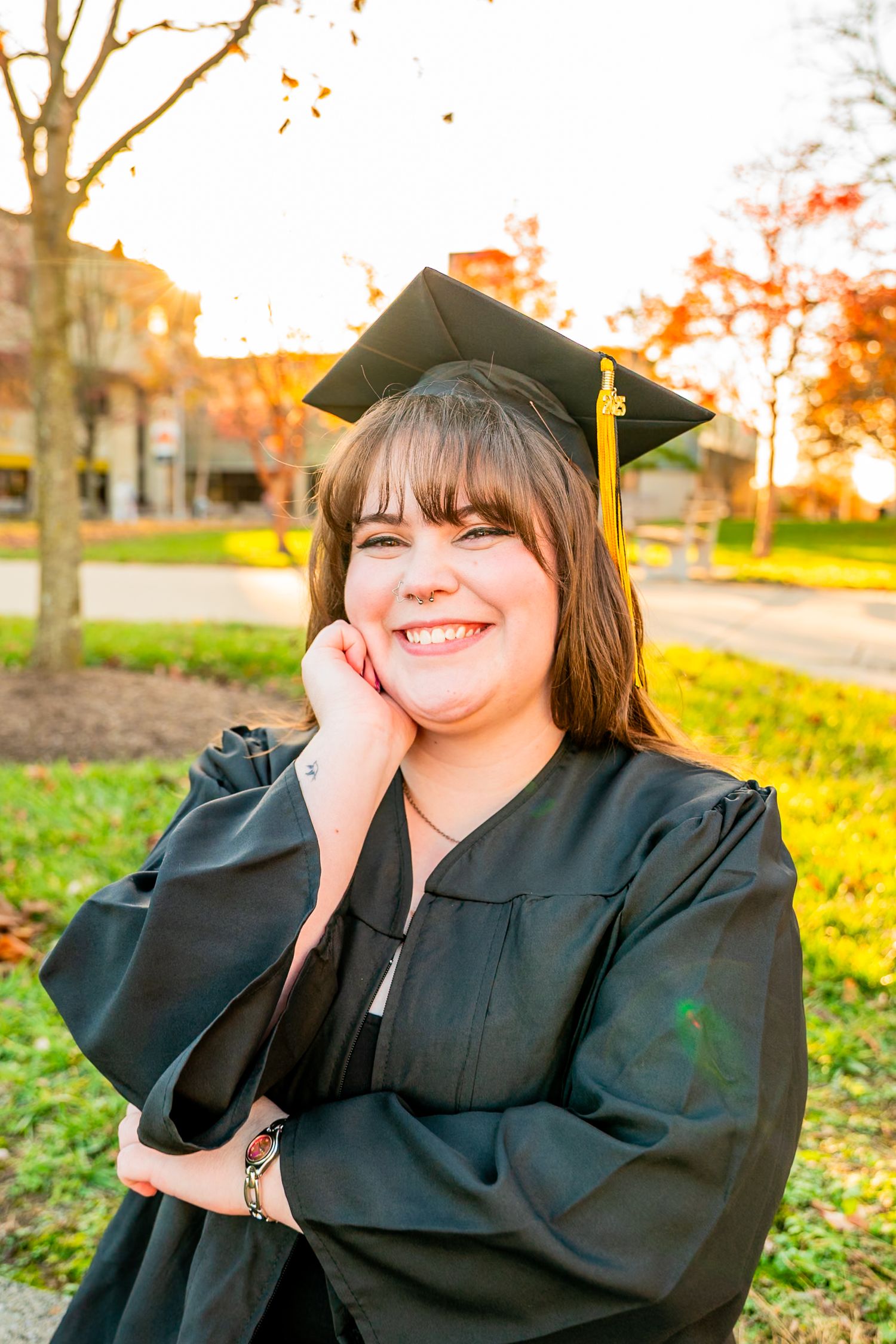 Graduate in black cap and gown smiles joyfully during autumn graduation photoshoot with colorful fall foliage in background.