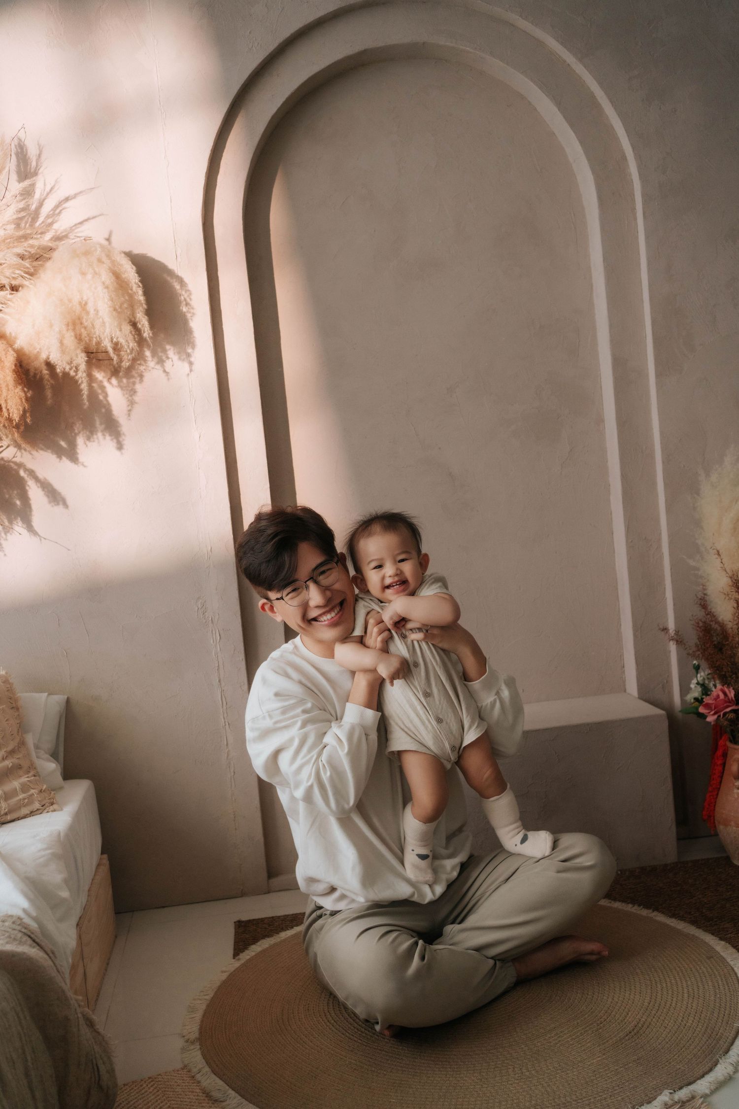 A young Asian man sits cross-legged on a round rug, smiling while holding a happy baby in a cozy neutral-toned room.一位年輕的亞洲男子盤腿坐在圓形地毯上，面帶微笑