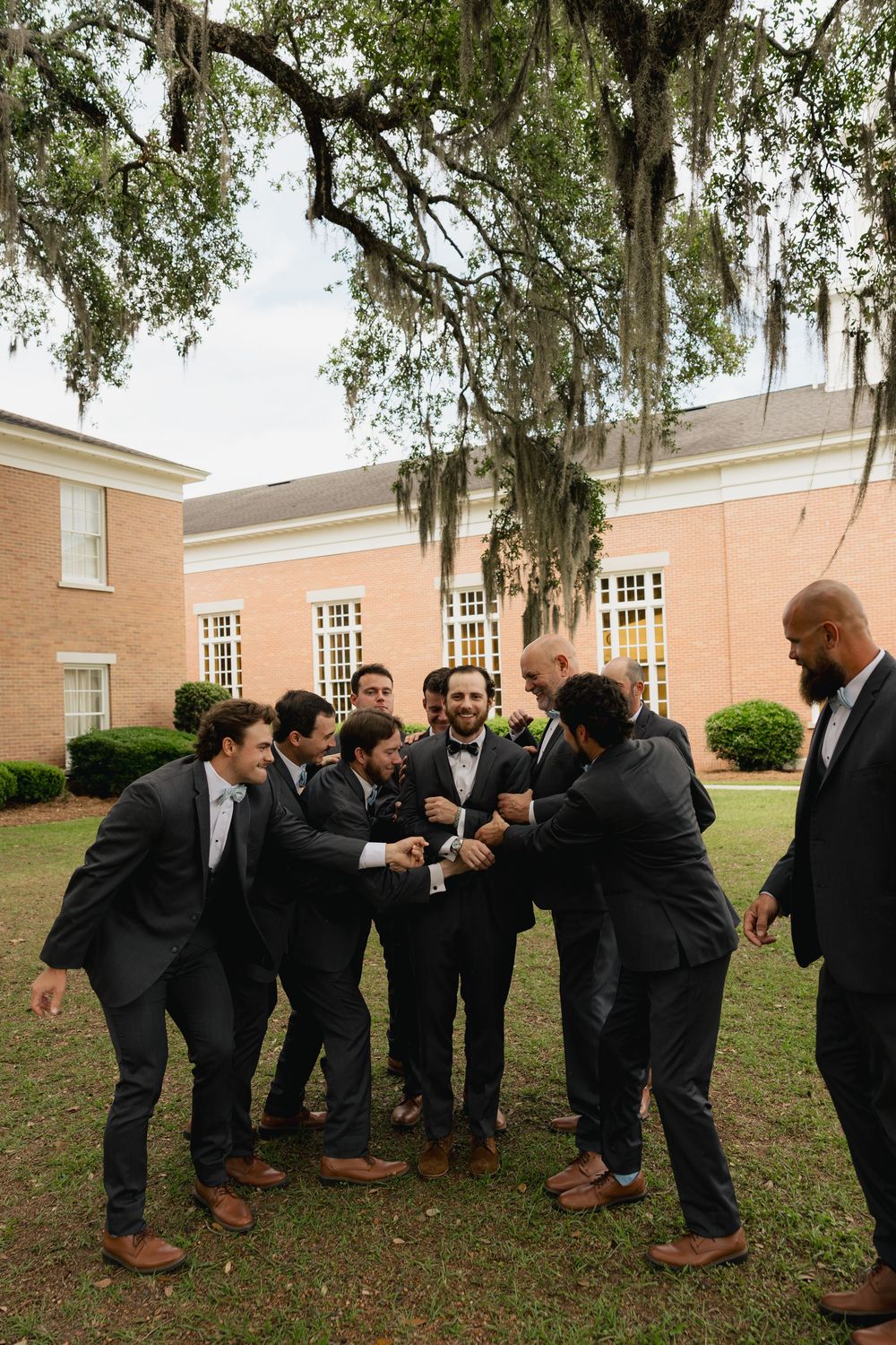 Groomsmen in dark suits celebrate with handshakes on a lawn outside a brick building with Spanish moss trees.