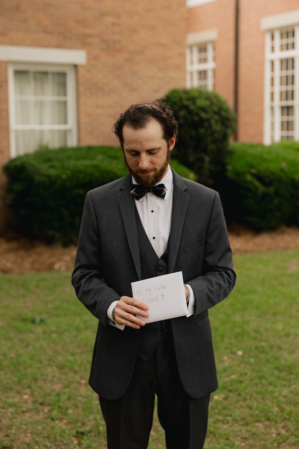 Groom in black tuxedo reading a wedding letter card outdoors near brick building and green shrubs.