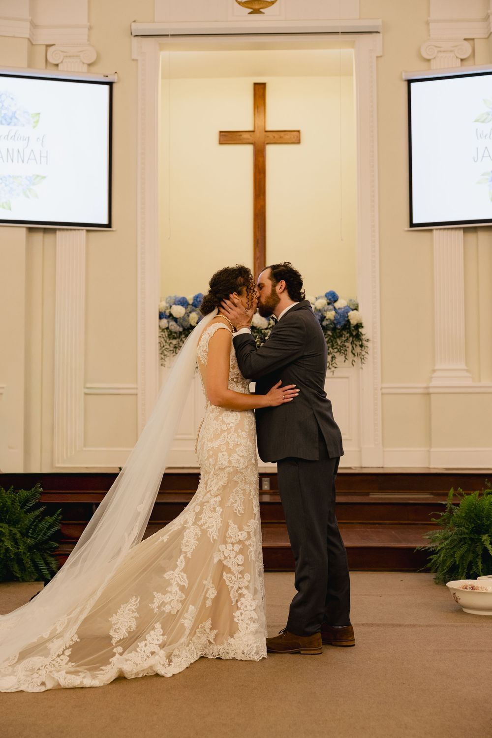 Bride and groom share first kiss at church altar with wooden cross and blue floral arrangements during wedding ceremony.