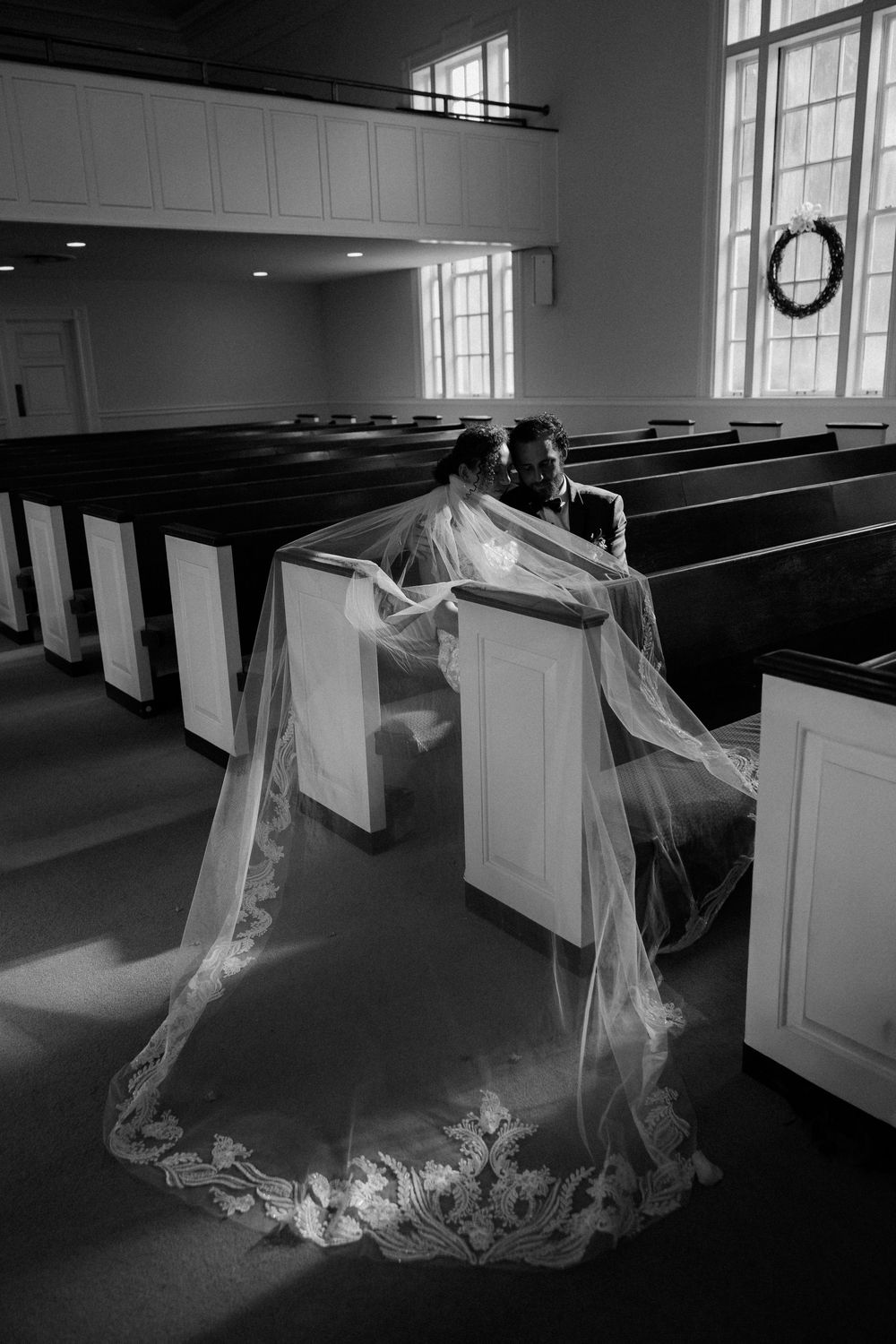 Black and white photo of bride and groom sitting in church pew, cathedral veil dramatically spread across aisle floor.