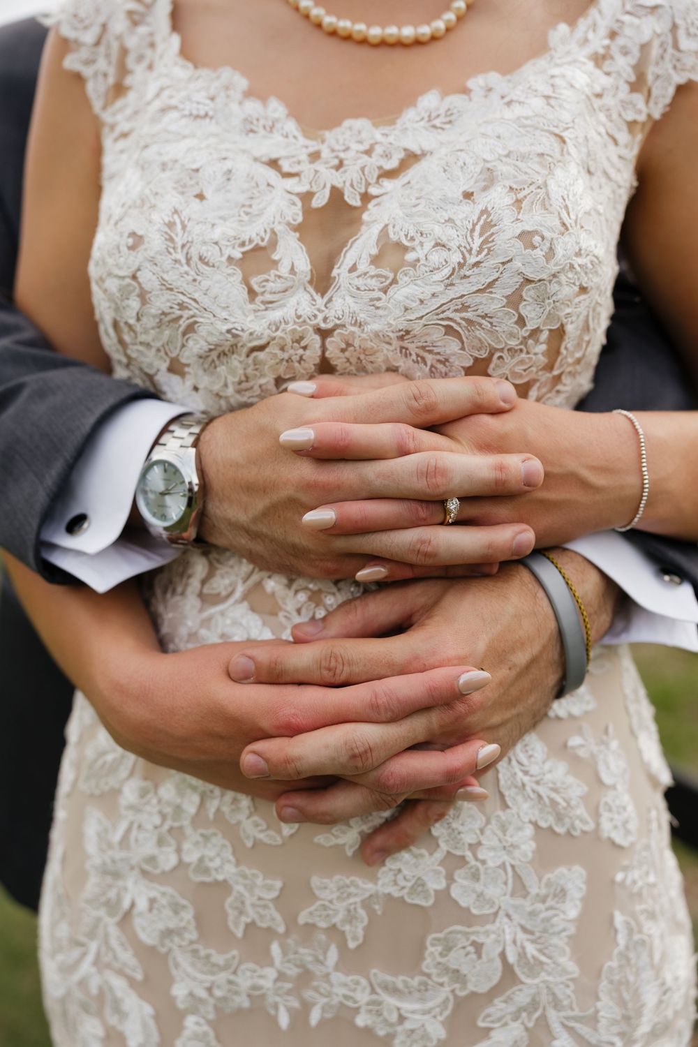 Bride and groom clasping hands together, showcasing wedding rings, a pearl necklace, and lace bridal gown.