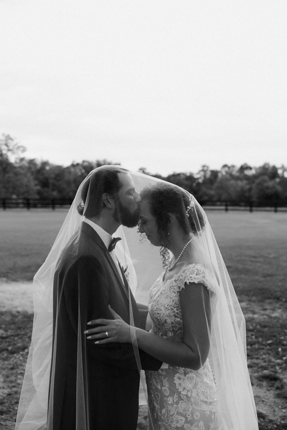 Black and white photo of bride and groom kissing under veil in a field.