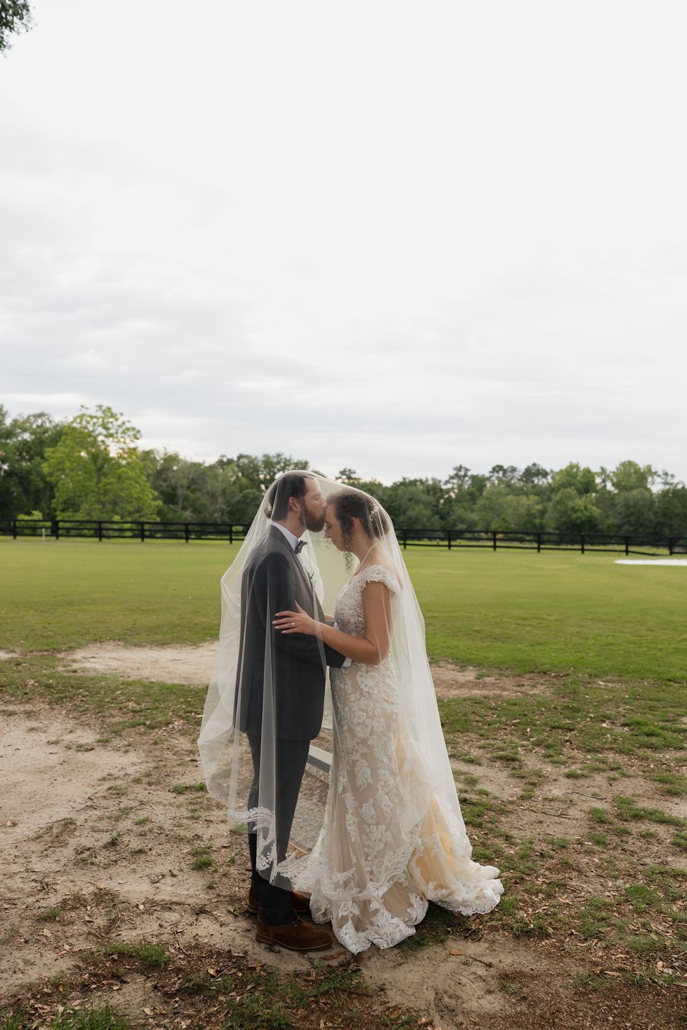 Bride and groom kissing under veil on a green field at outdoor wedding.