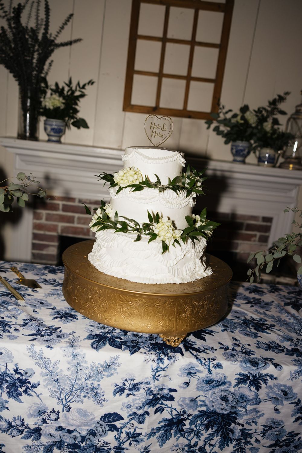 Three-tier white wedding cake with greenery and flowers on gold stand, blue floral tablecloth, brick fireplace backdrop.