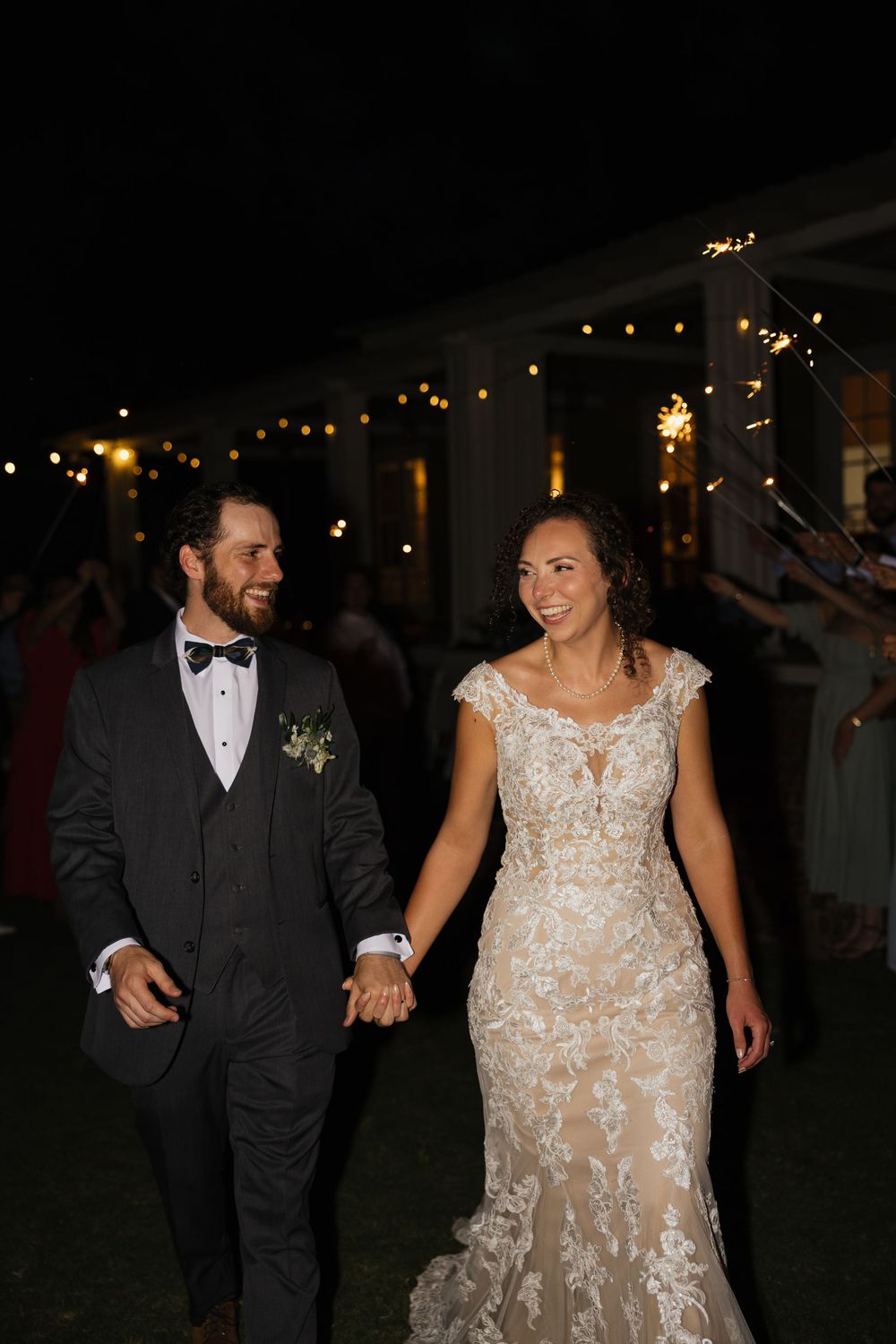 A smiling bride and groom walk hand in hand at night surrounded by sparklers during their wedding reception.
