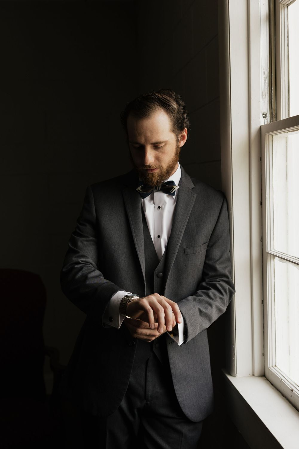 A groom in a gray tuxedo and bow tie checks his watch by a window in soft natural light.