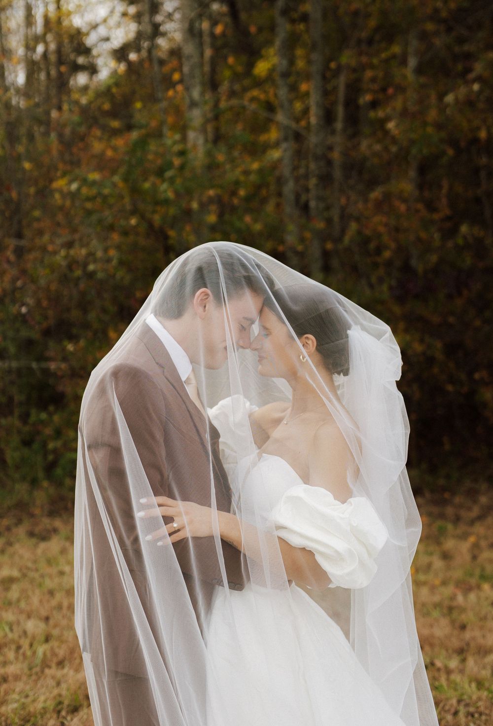 A romantic wedding photo showing a couple sharing an intimate moment under a delicate white veil in an autumn setting.