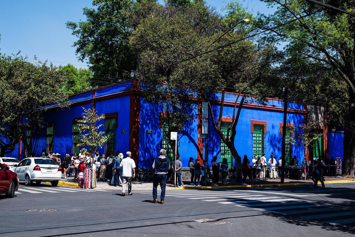 Vibrant blue building exterior with people walking on sidewalk near trees and parked cars in urban setting.